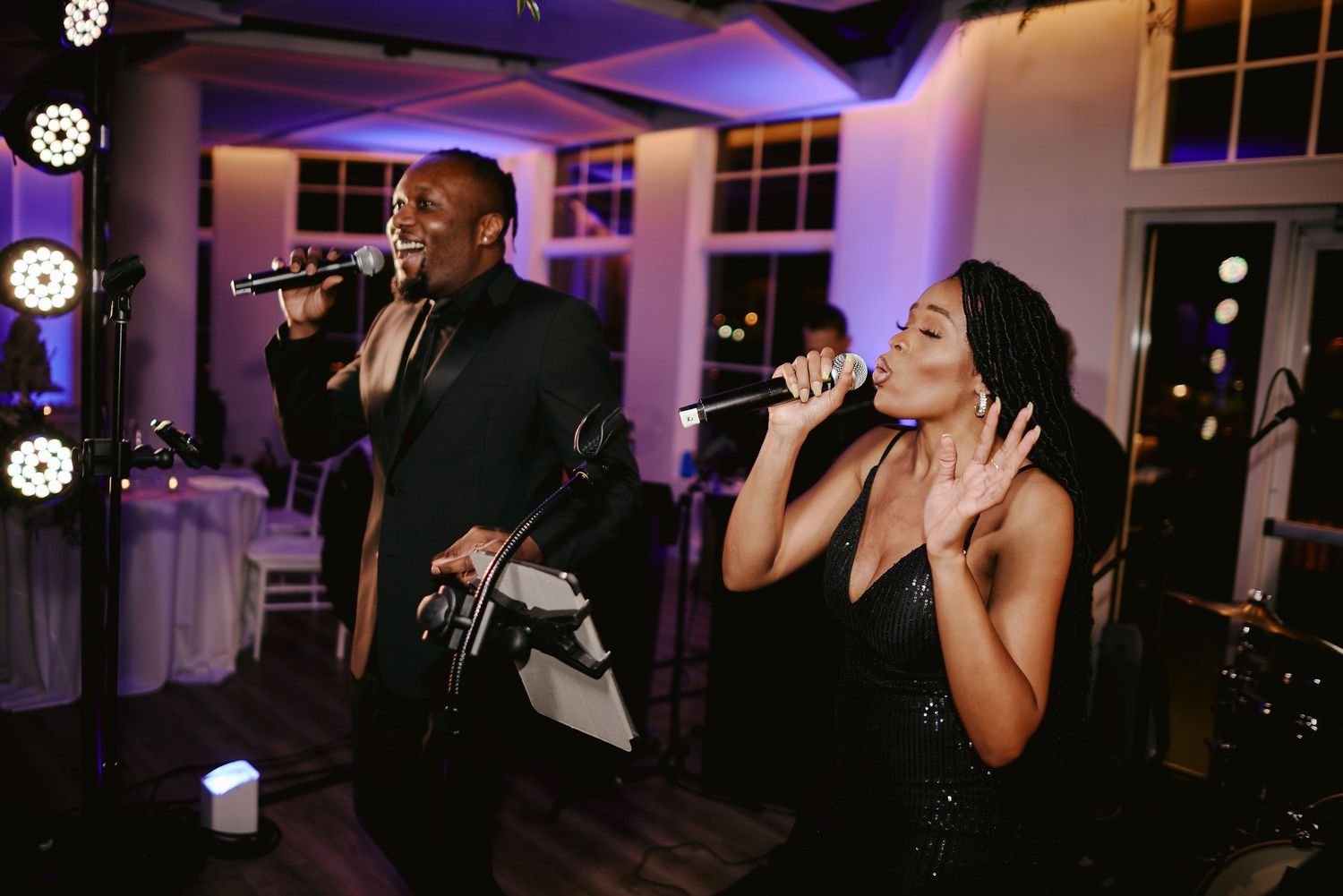 Two performers sing with microphones at an evening reception venue illuminated by purple lighting.