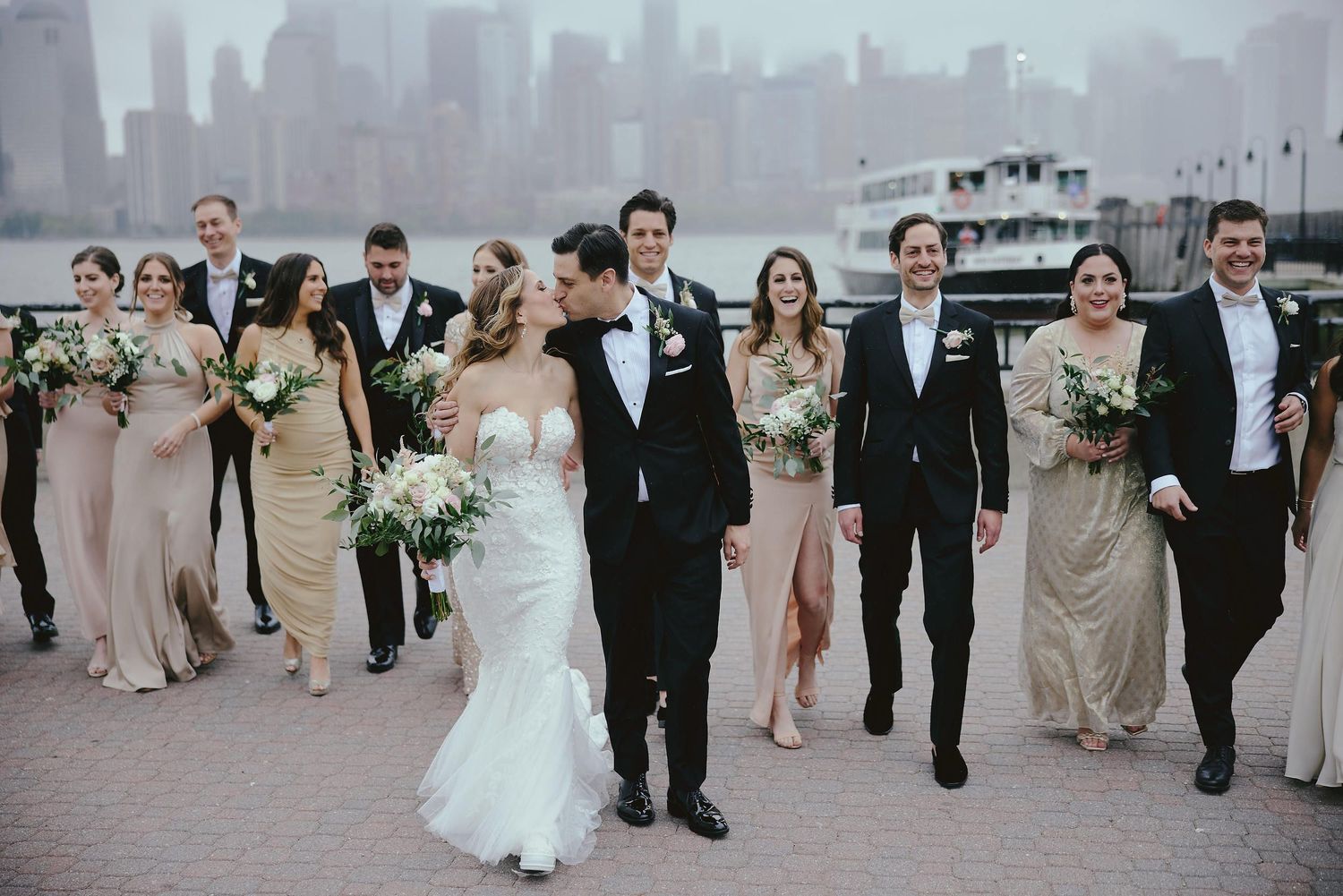 Wedding party walks along foggy city waterfront with bride in white gown and attendants in neutral dresses.