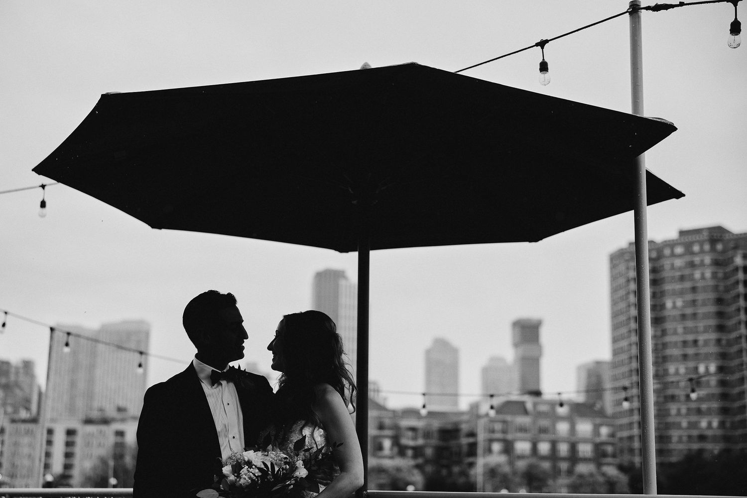 A silhouetted couple shares an intimate moment under a dark umbrella against a cityscape backdrop.