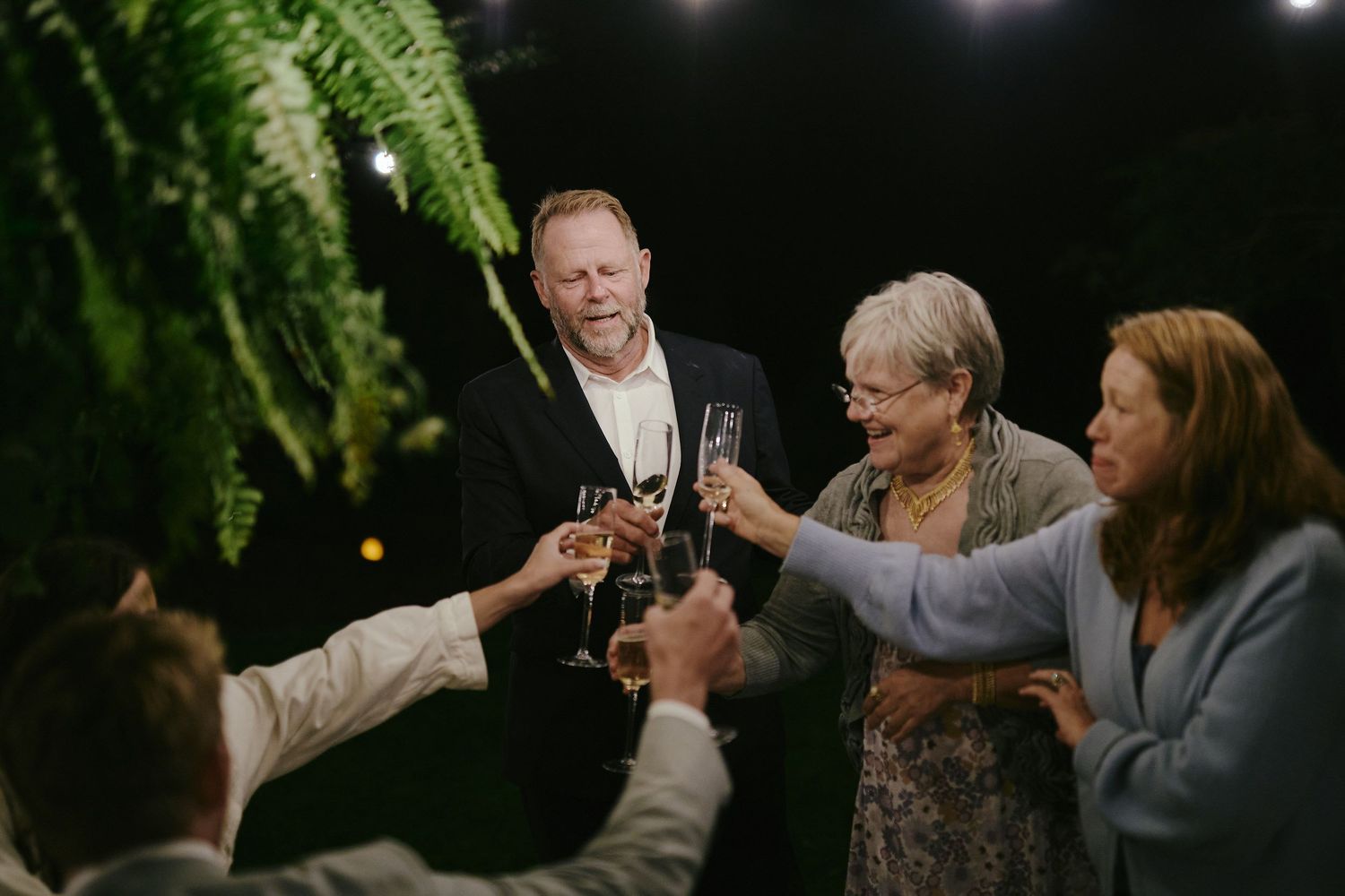 A group of people raising champagne glasses for a celebratory toast under hanging ferns and evening lights.