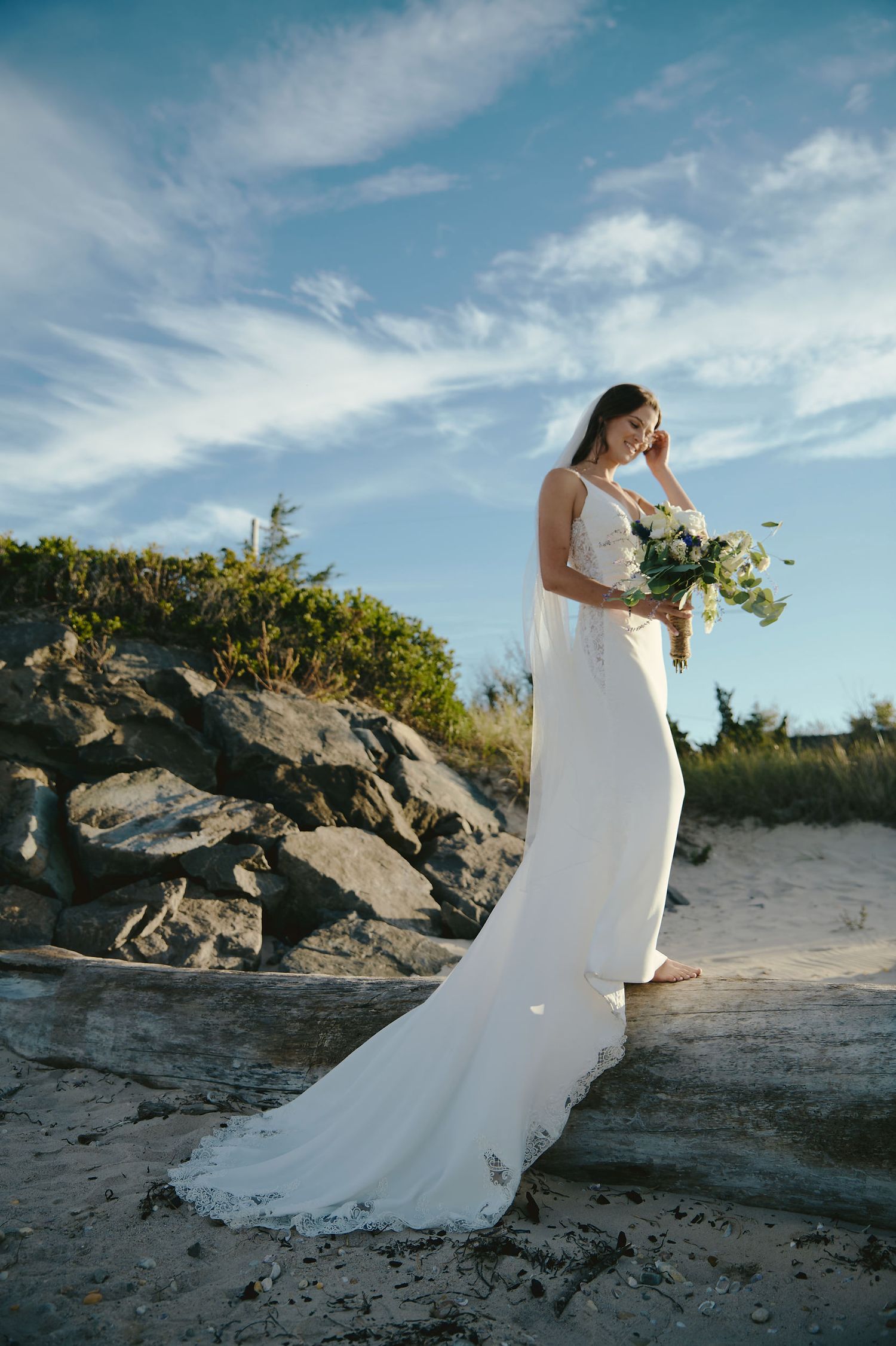 A bride in a flowing white gown holds a bouquet on a rocky beach with a dramatic blue sky and wispy clouds.