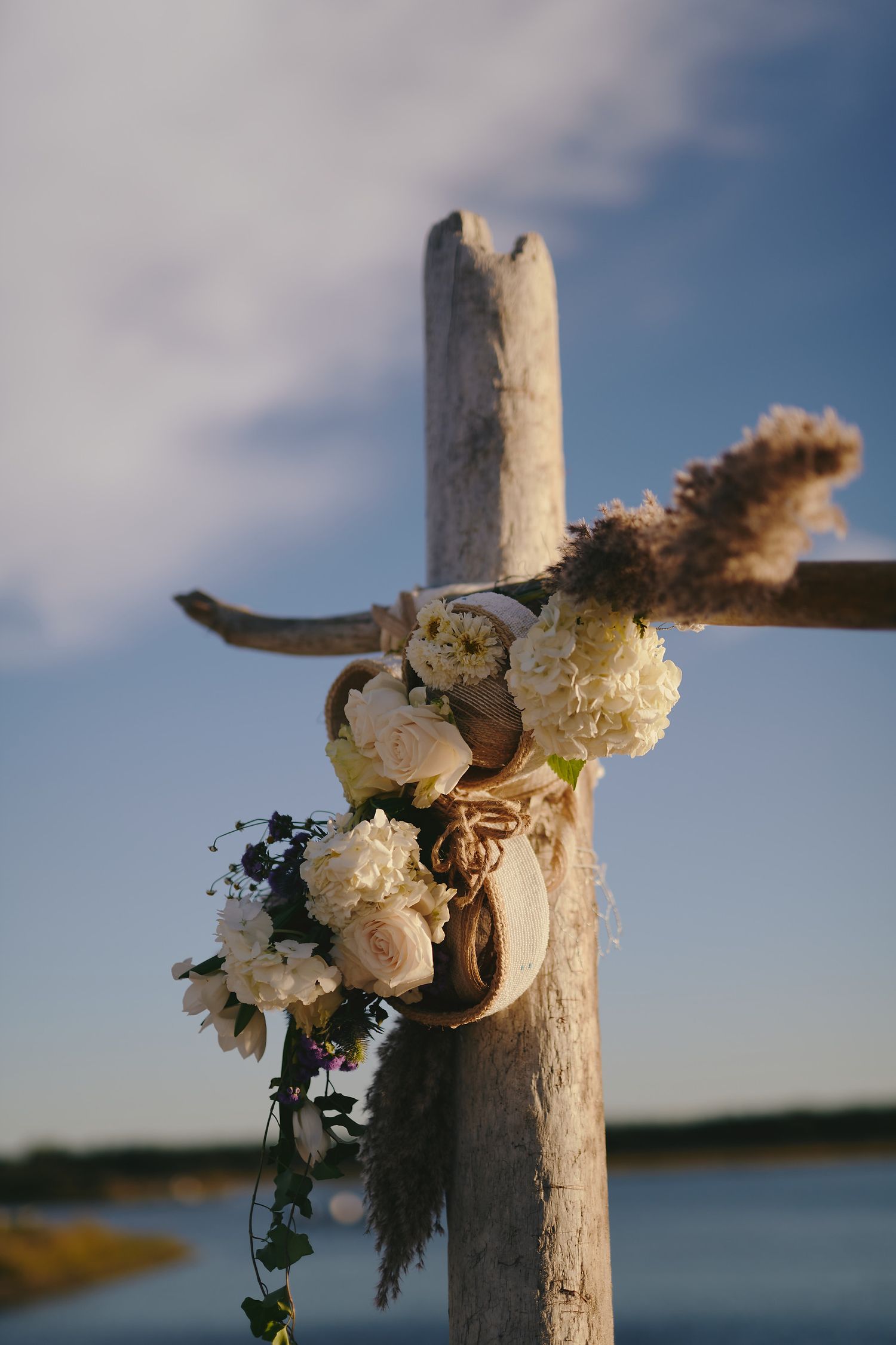 A wooden cross adorned with white roses and floral arrangements stands against a serene waterfront backdrop.