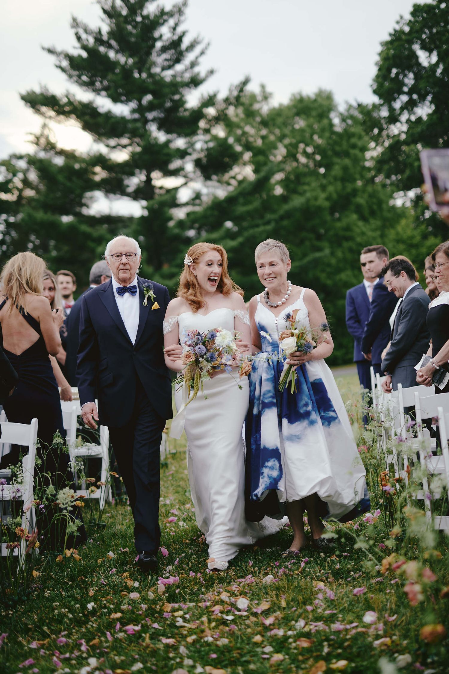 White chairs line an outdoor wedding aisle with scattered petals and evergreen trees in the background.