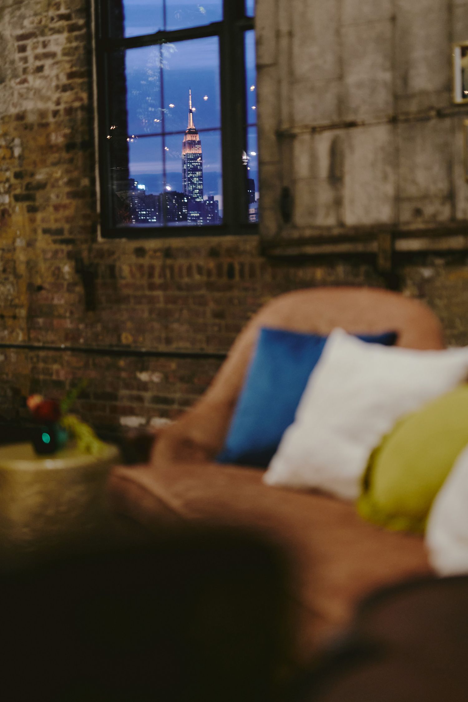 A cozy apartment living room with colorful throw pillows and a window view of the Empire State Building at night.