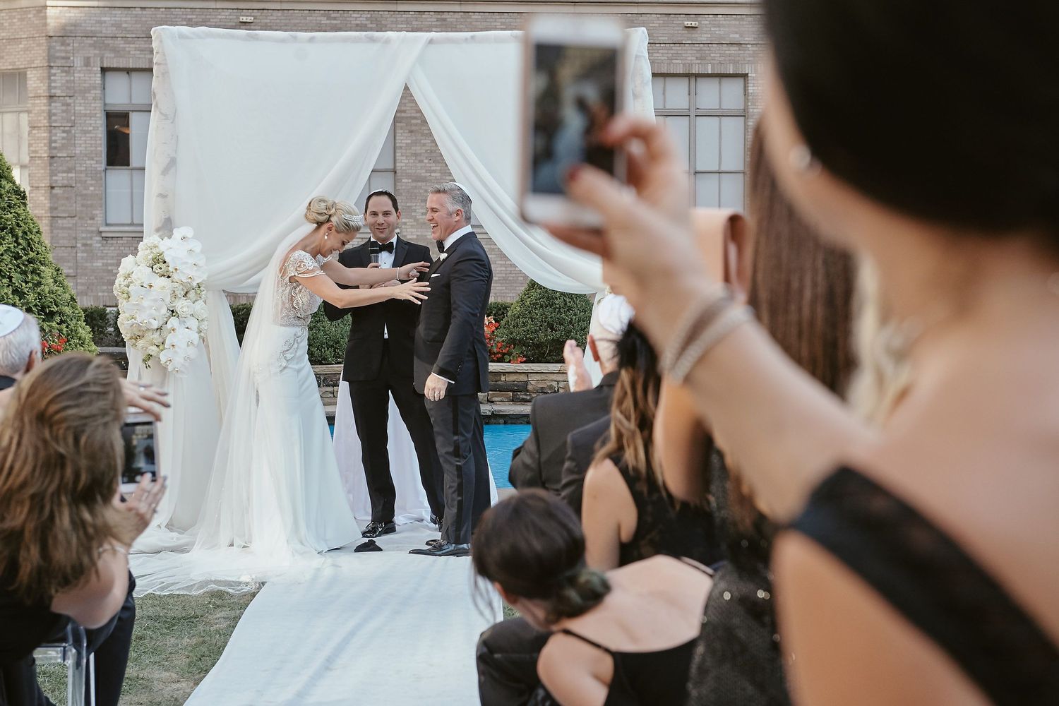 Wedding ceremony taking place outdoors under white draped arch with guests watching and taking photos.