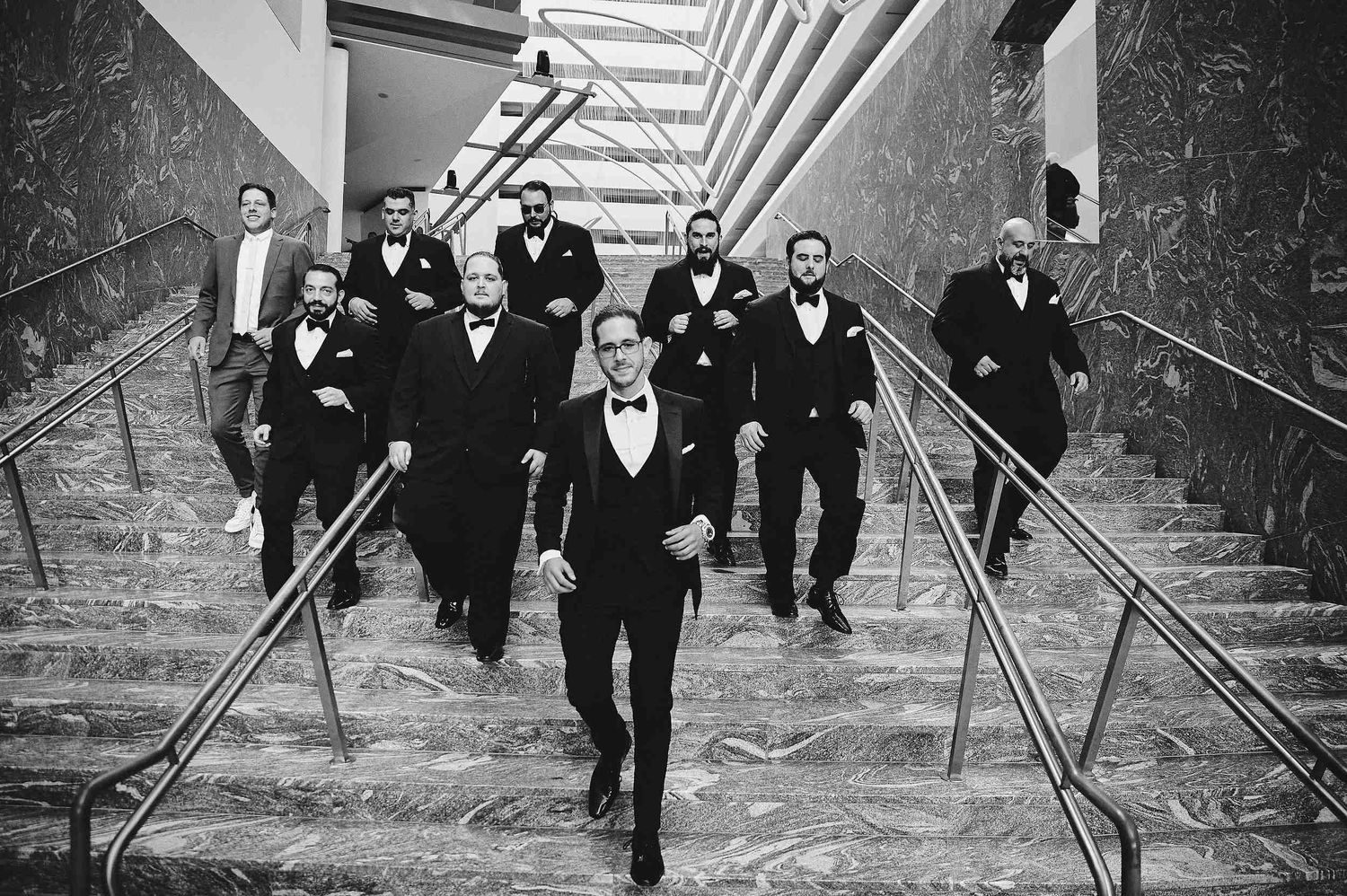 Black and white photo of wedding party in formal attire descending grand marble staircase with elegant handrails.