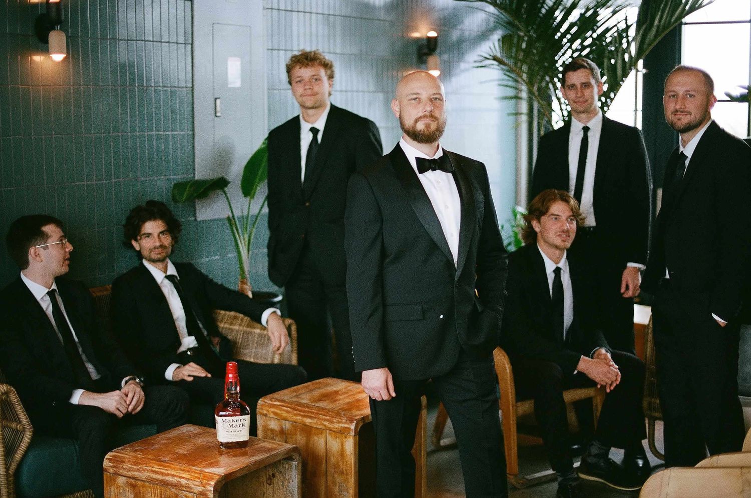 Groomsmen in formal attire gather around wooden furniture with champagne bottles at an elegant indoor venue.