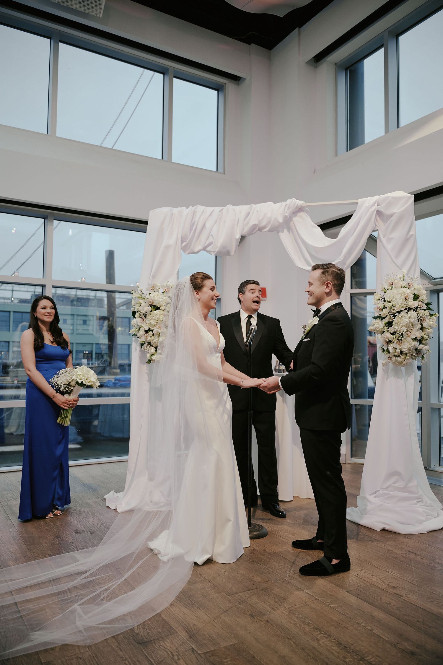 Indoor wedding ceremony takes place under white fabric arch with floral arrangements in modern venue with city views.