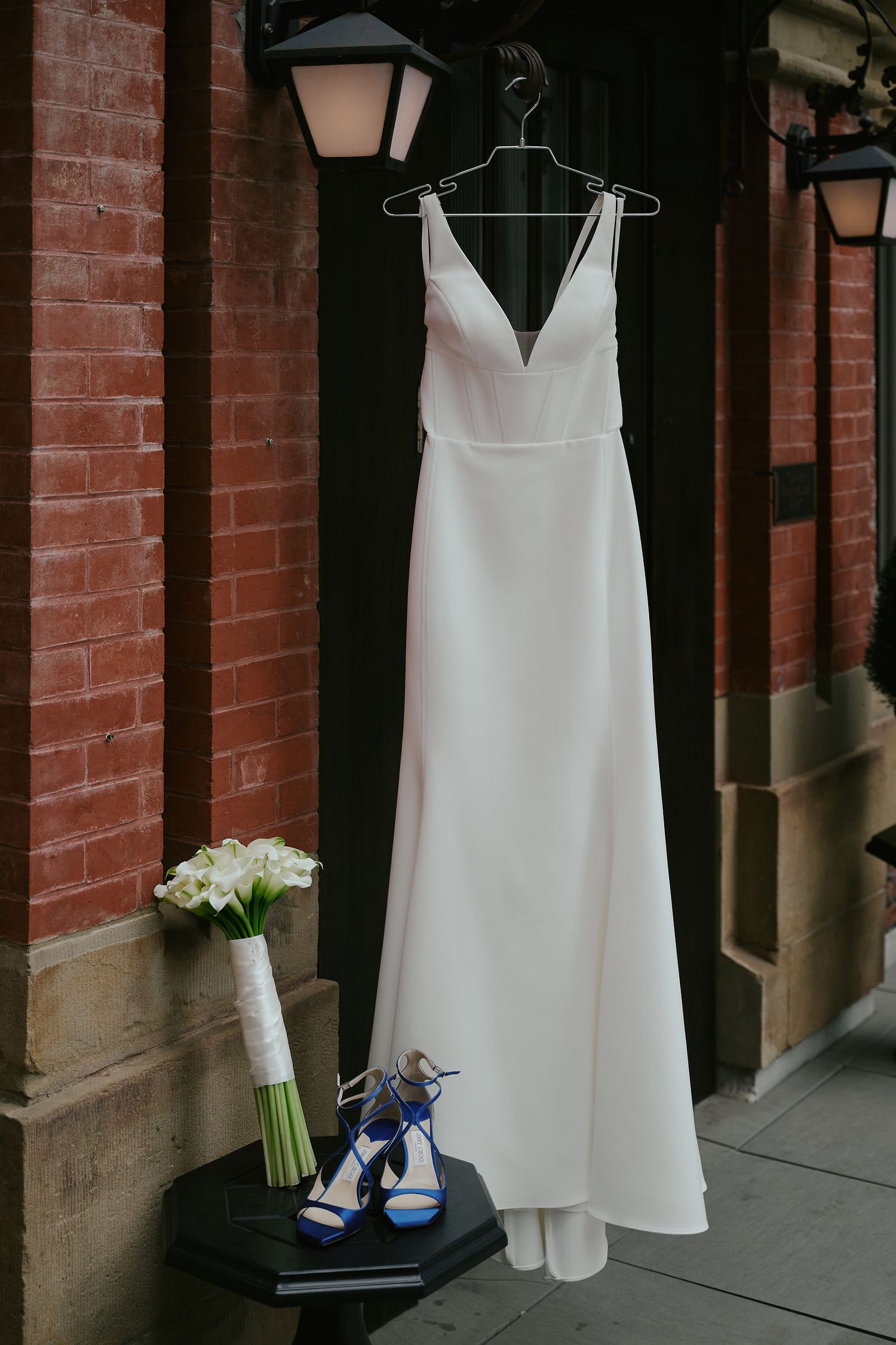 Elegant white wedding dress hanging on brick wall with blue heels and white bouquet displayed below.