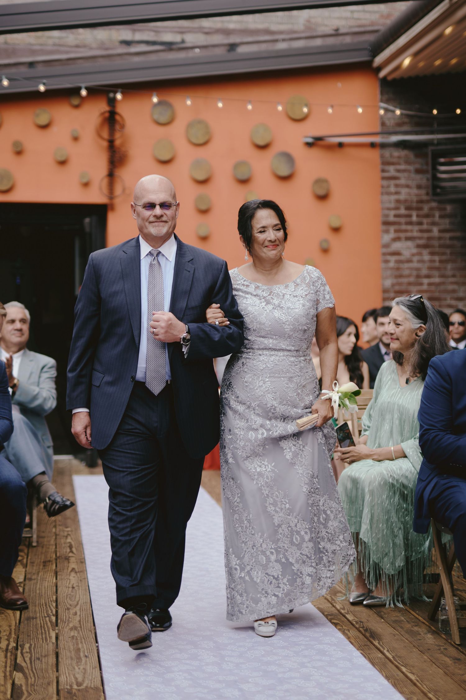 A couple walks arm-in-arm down a rustic wooden aisle with an orange wall adorned with circular decorations behind them.