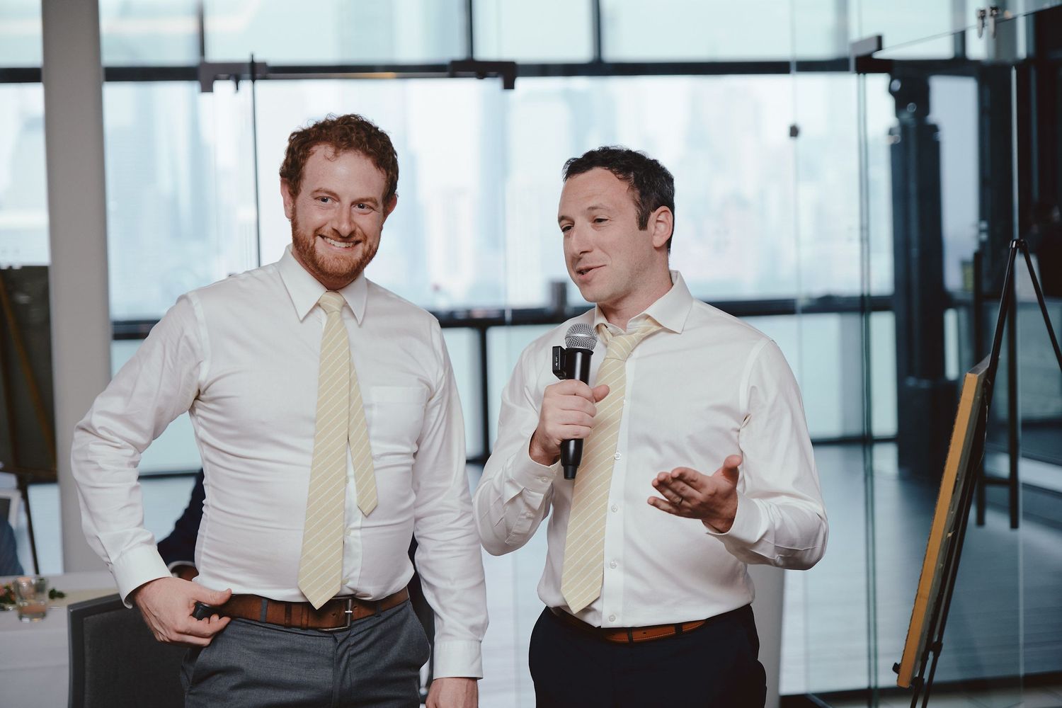Two groomsmen in white dress shirts share a laugh while giving wedding speeches at an indoor venue.