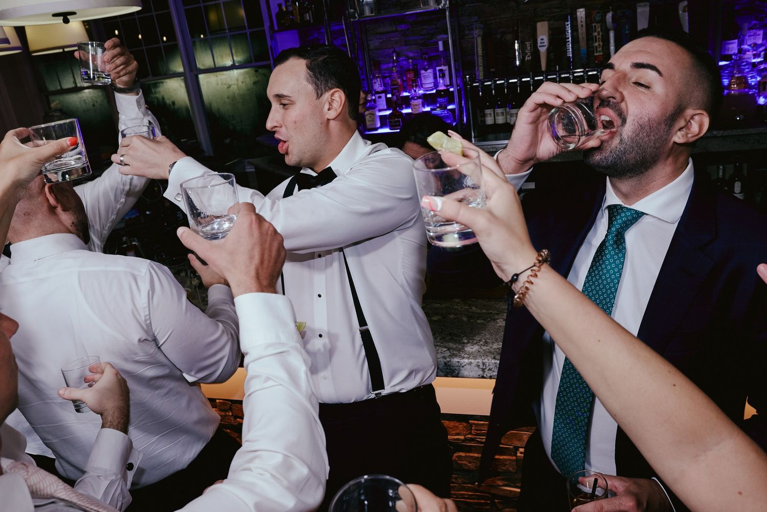 A group of people at a bar enthusiastically taking shots of drinks together in a nightclub setting with purple lighting.