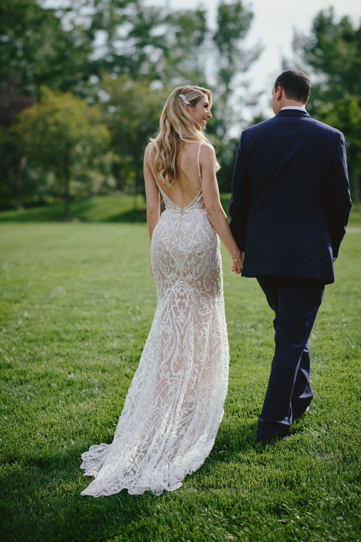 A couple in wedding attire walk on a lush green lawn with the bride wearing an intricate lace backless gown.