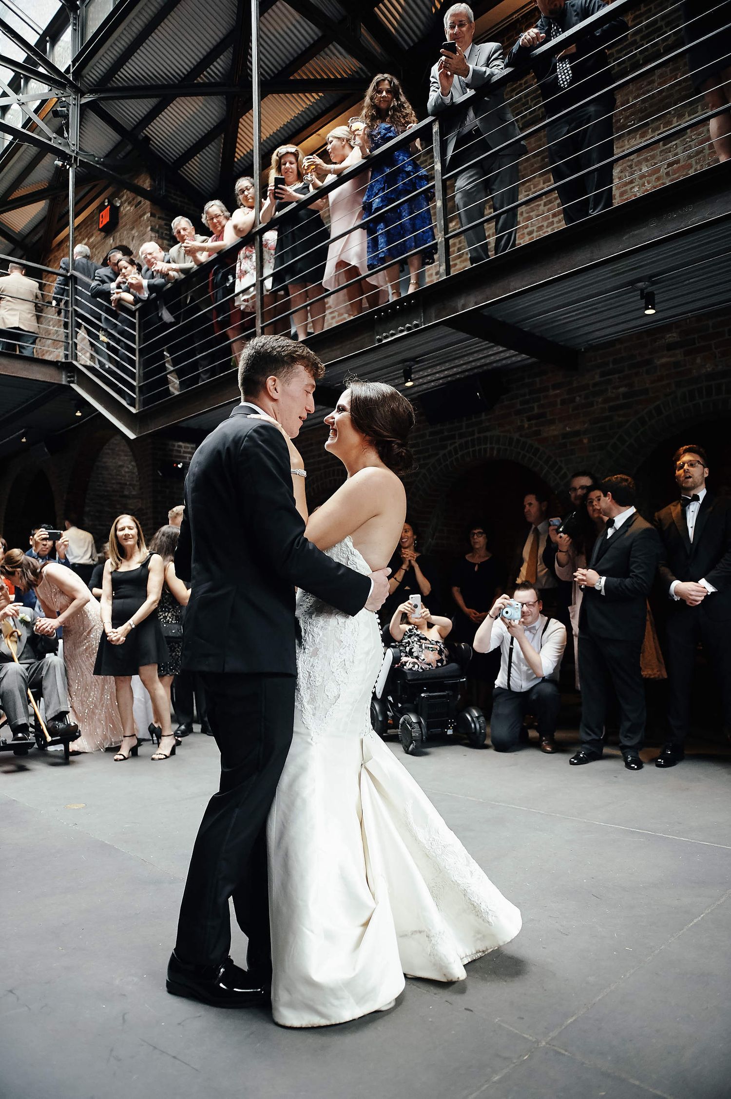 Wedding guests watch from above as newlyweds share their first dance in an industrial-style venue.
