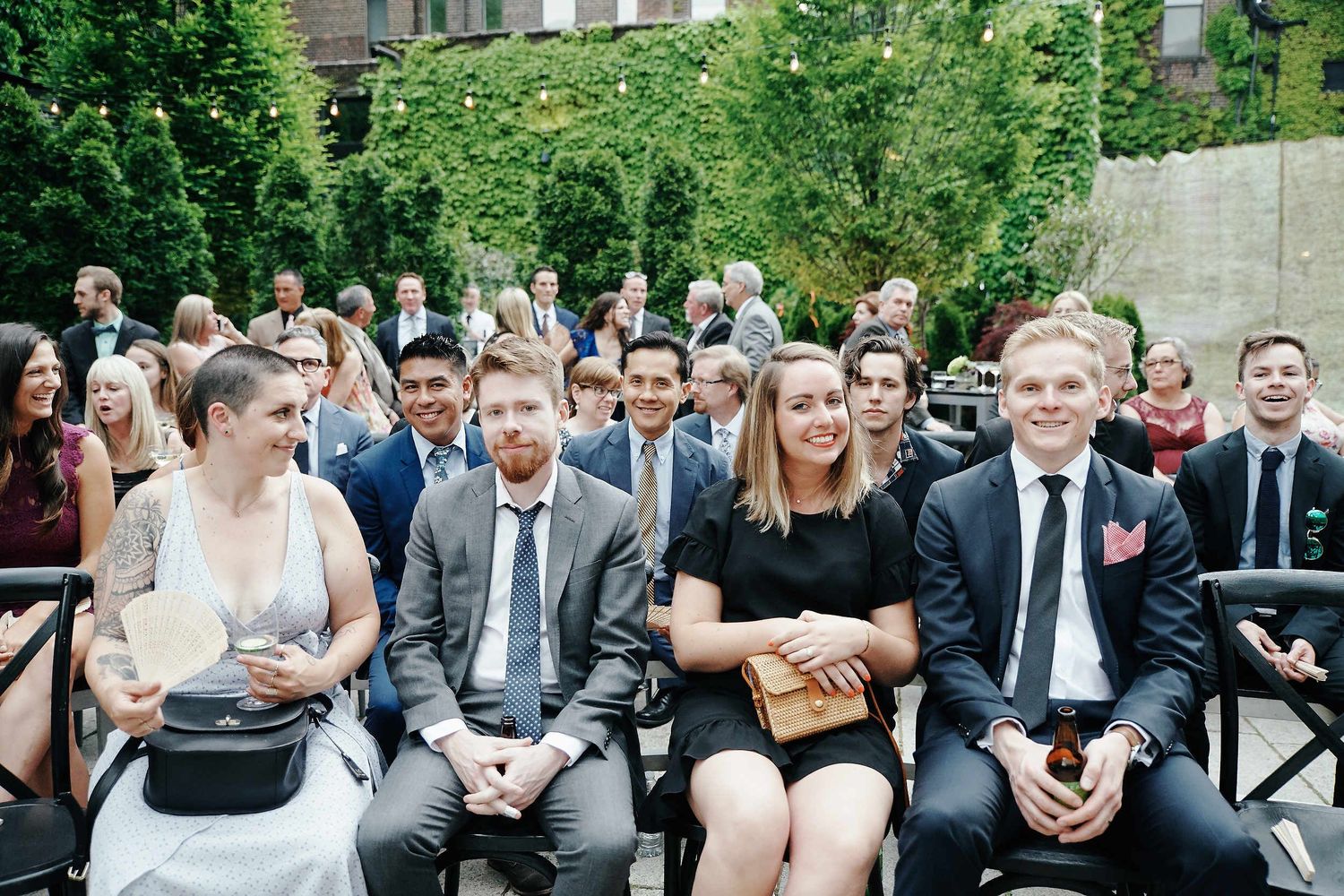 Wedding guests seated in rows outdoors at a garden ceremony with lush greenery in the background.