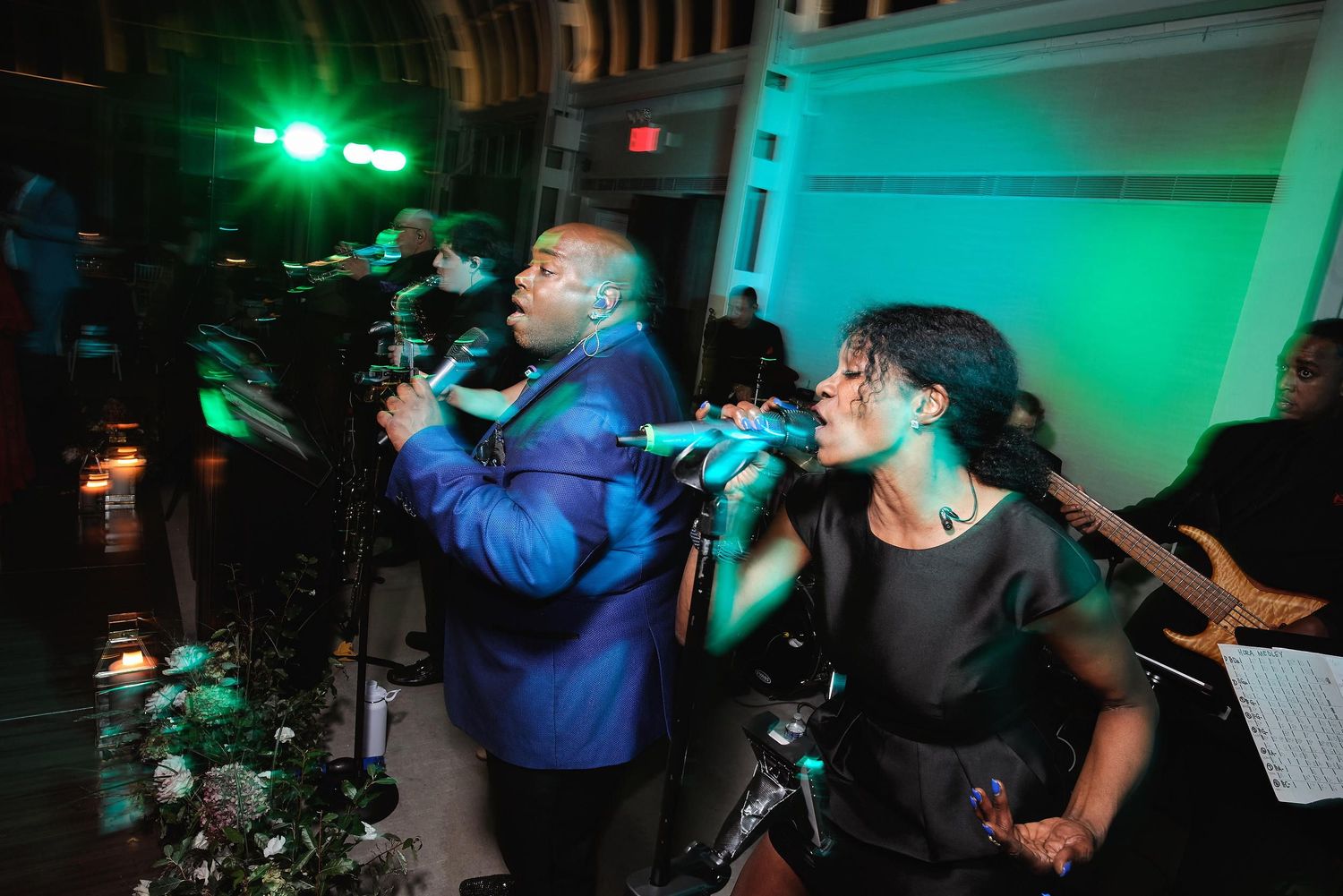 Band members perform energetically with microphones under dramatic green stage lighting at wedding reception.
