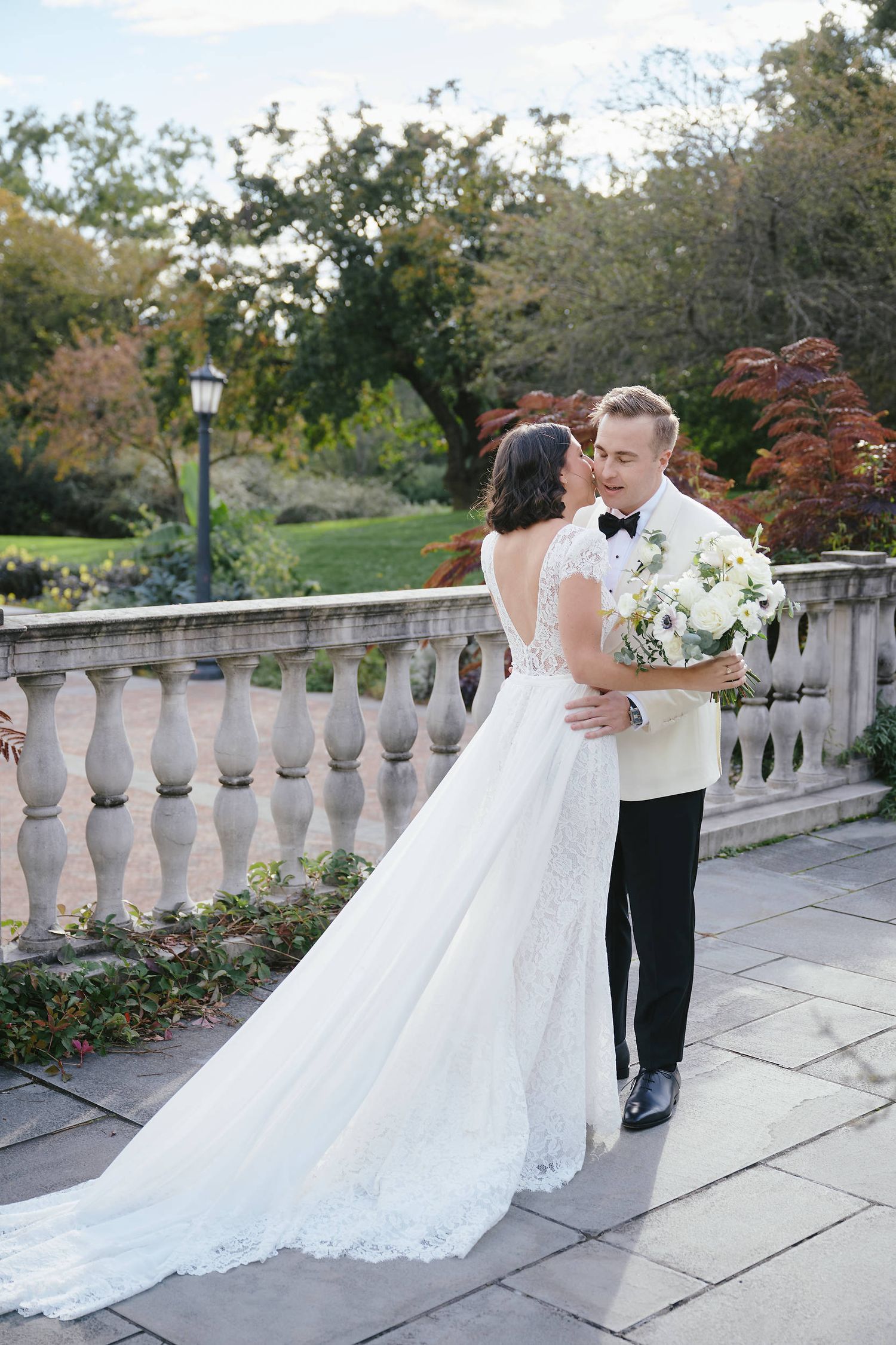 Couple in wedding attire embrace on stone balcony terrace with white floral bouquet and autumn foliage in background.