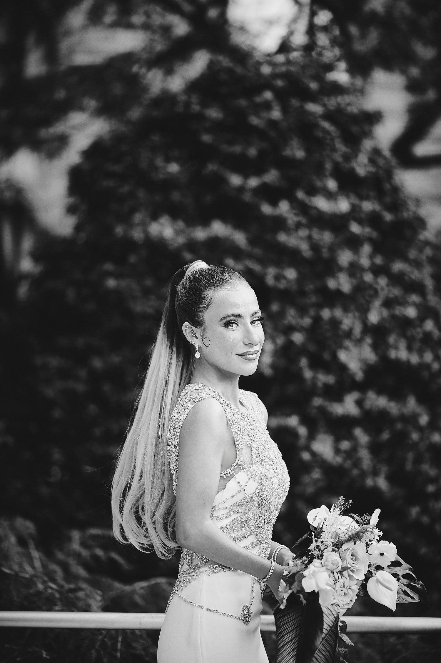 A bride in a lace wedding dress poses with her bouquet in a black and white outdoor portrait.
