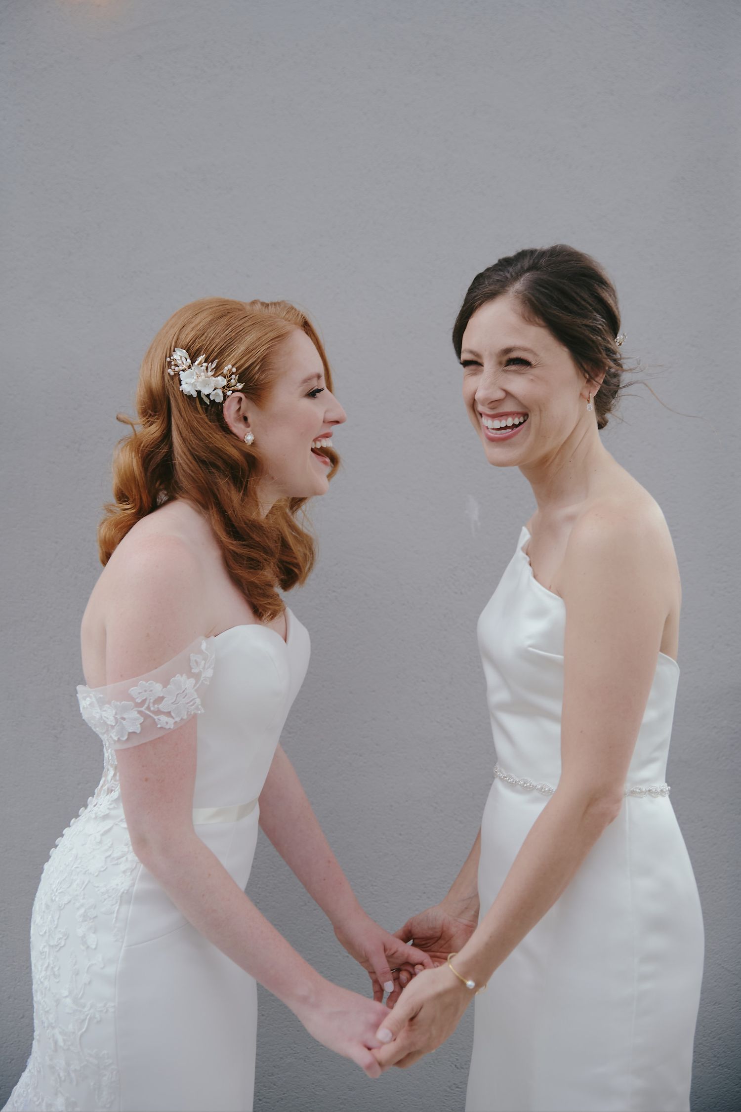 Two brides in white wedding dresses share a joyful moment laughing together against a grey background.