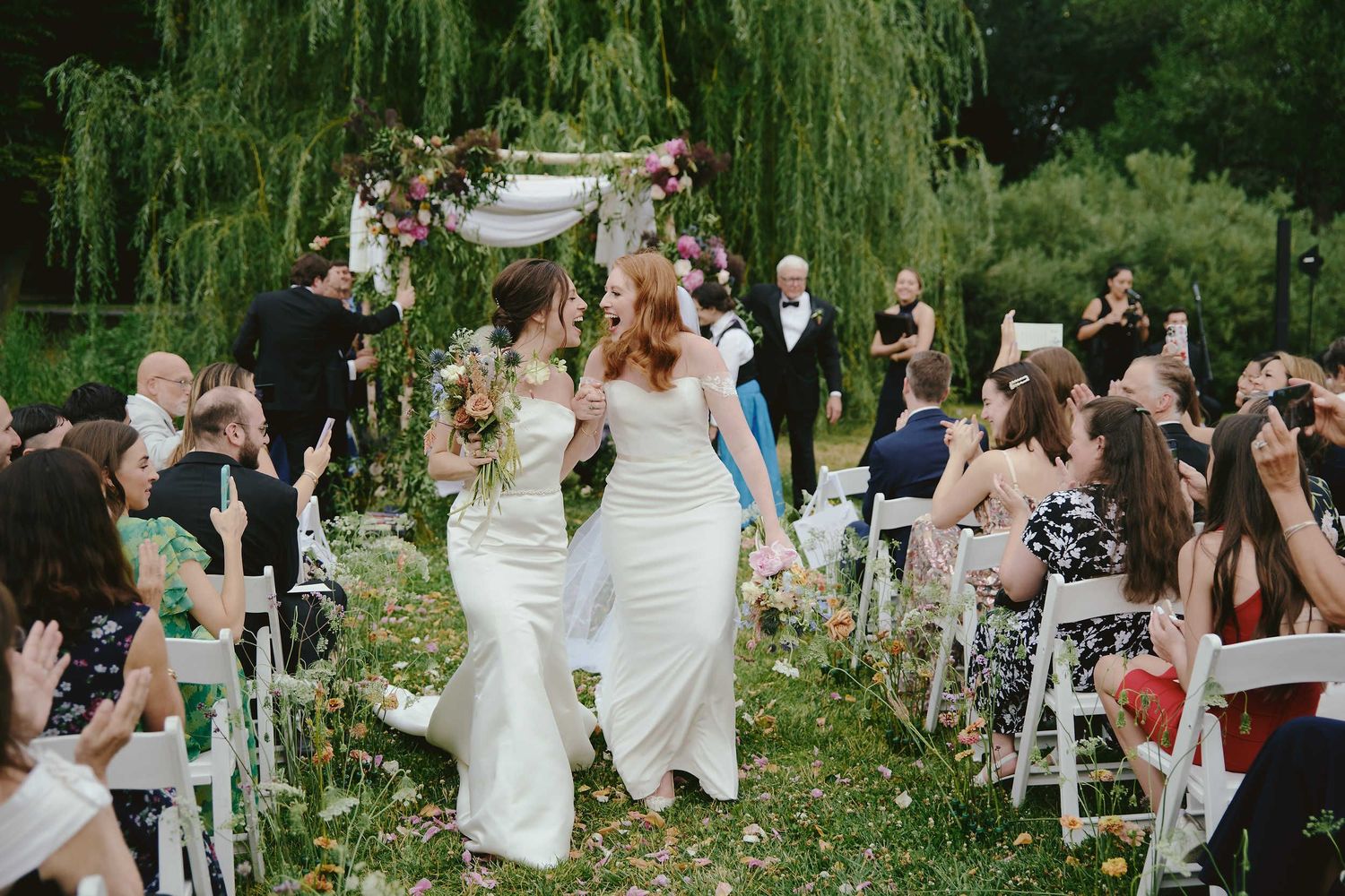 Two brides in white wedding dresses walk down the aisle together at an outdoor garden ceremony with weeping willows.