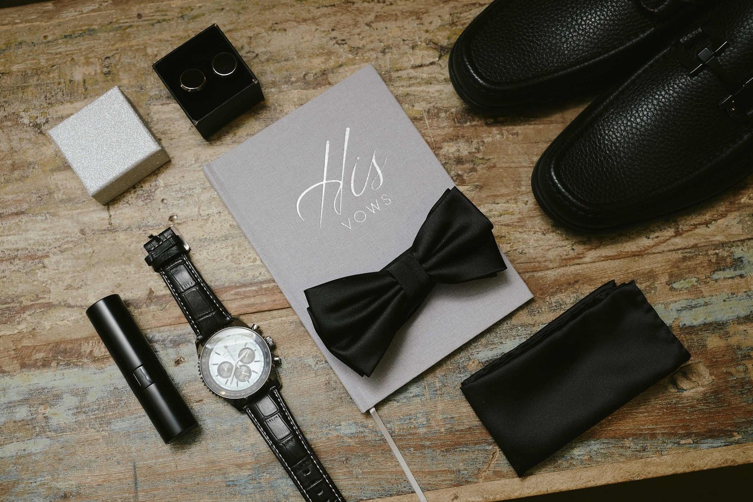 Wedding accessories including black bow tie, watch, dress shoes and groom's details card arranged on wooden surface.