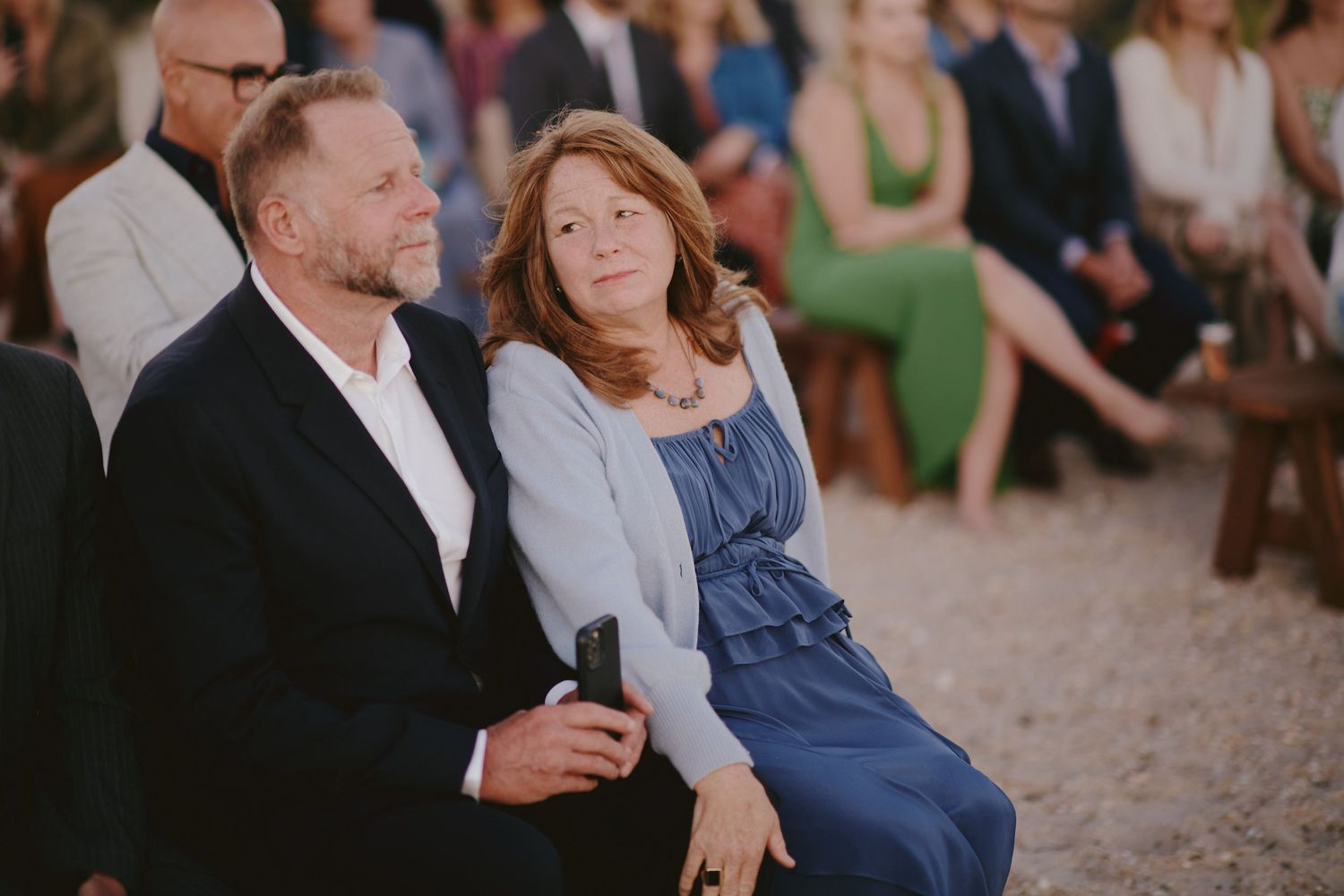 A couple sits together at an outdoor event, the woman wearing a blue dress while leaning affectionately toward her partner.