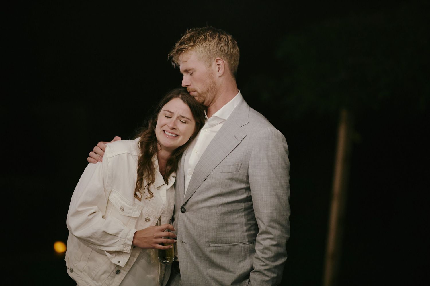 A couple in light colored formal wear embrace and share a tender moment against a dark background at night.