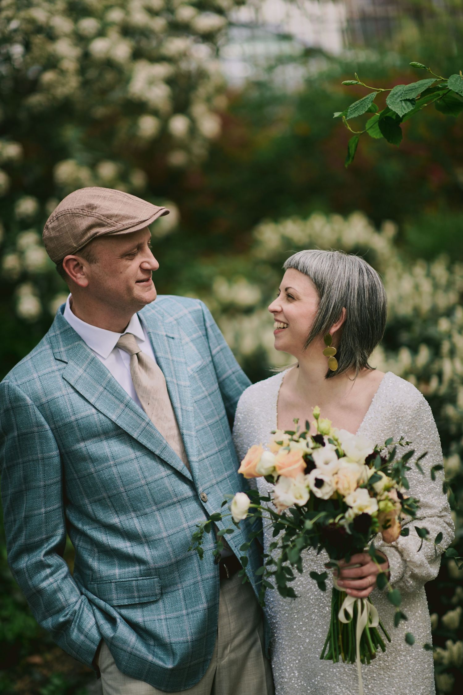 Couple in vintage-inspired wedding attire share loving glance while holding floral bouquet.