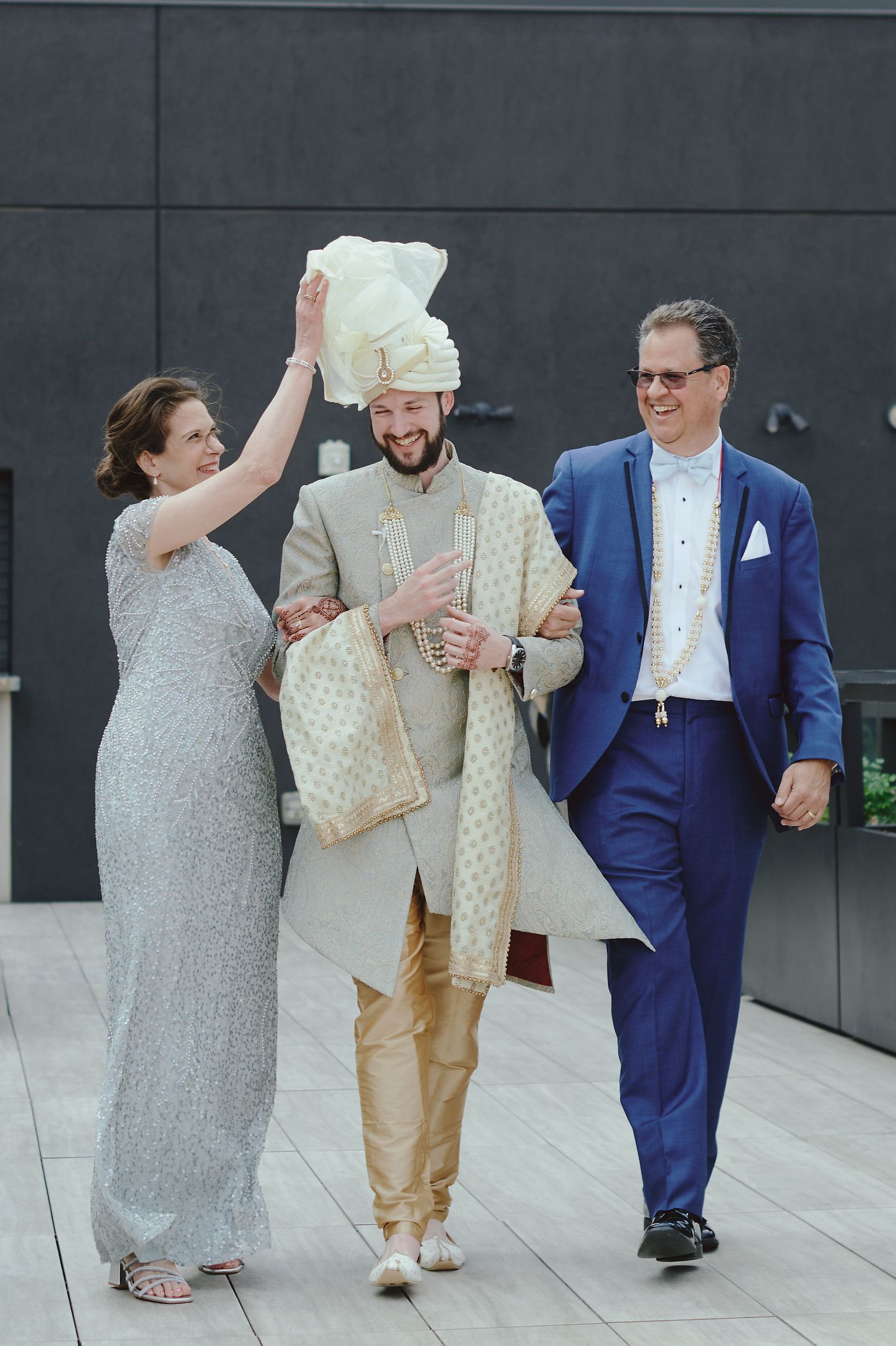 Three people in formal attire walk together on a paved area, with one wearing traditional ethnic clothing in cream tones.