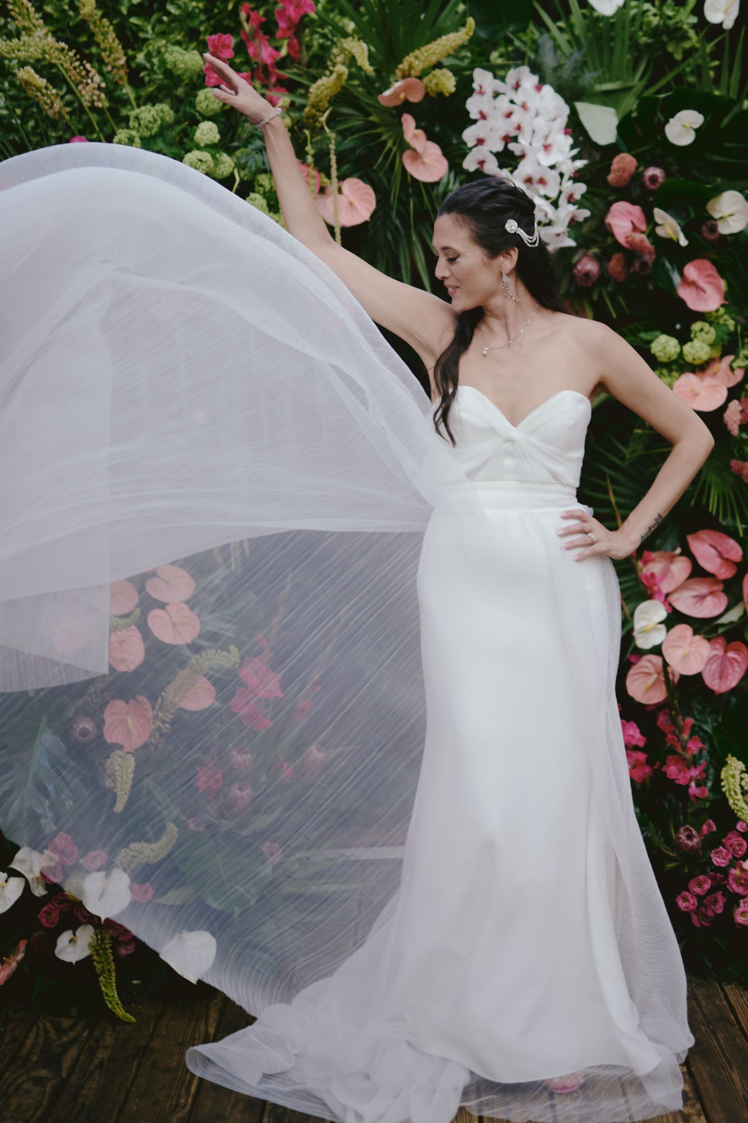 Elegant bride in flowing white gown poses dramatically with veil against floral backdrop of pink flowers and greenery.