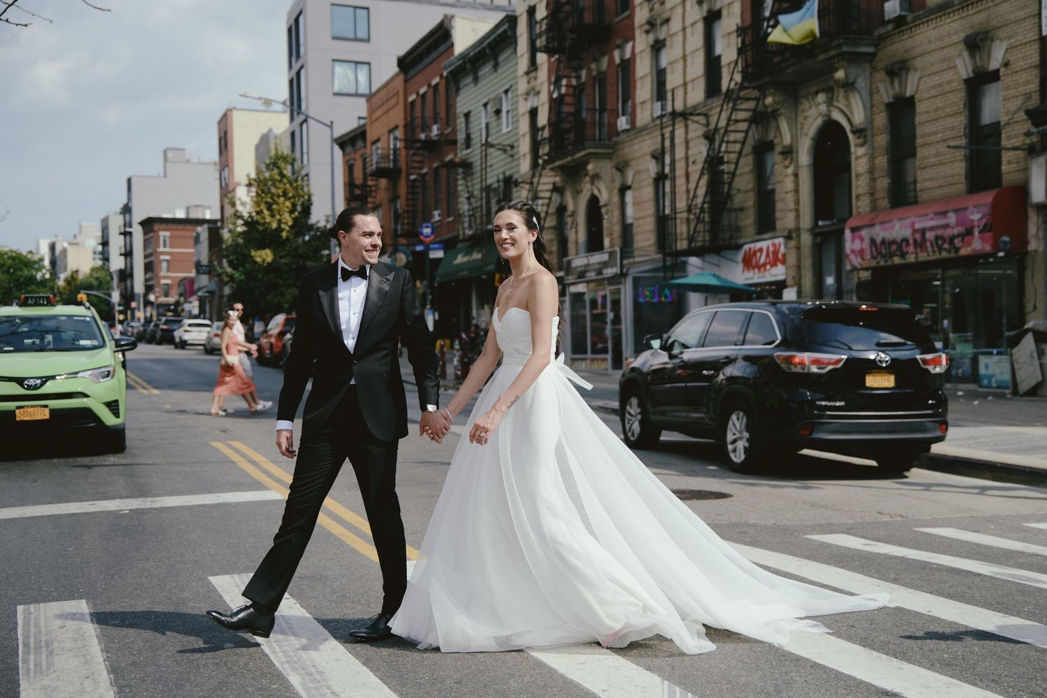 A couple in formal wedding attire crossing an urban street with historic buildings and cars in the background.