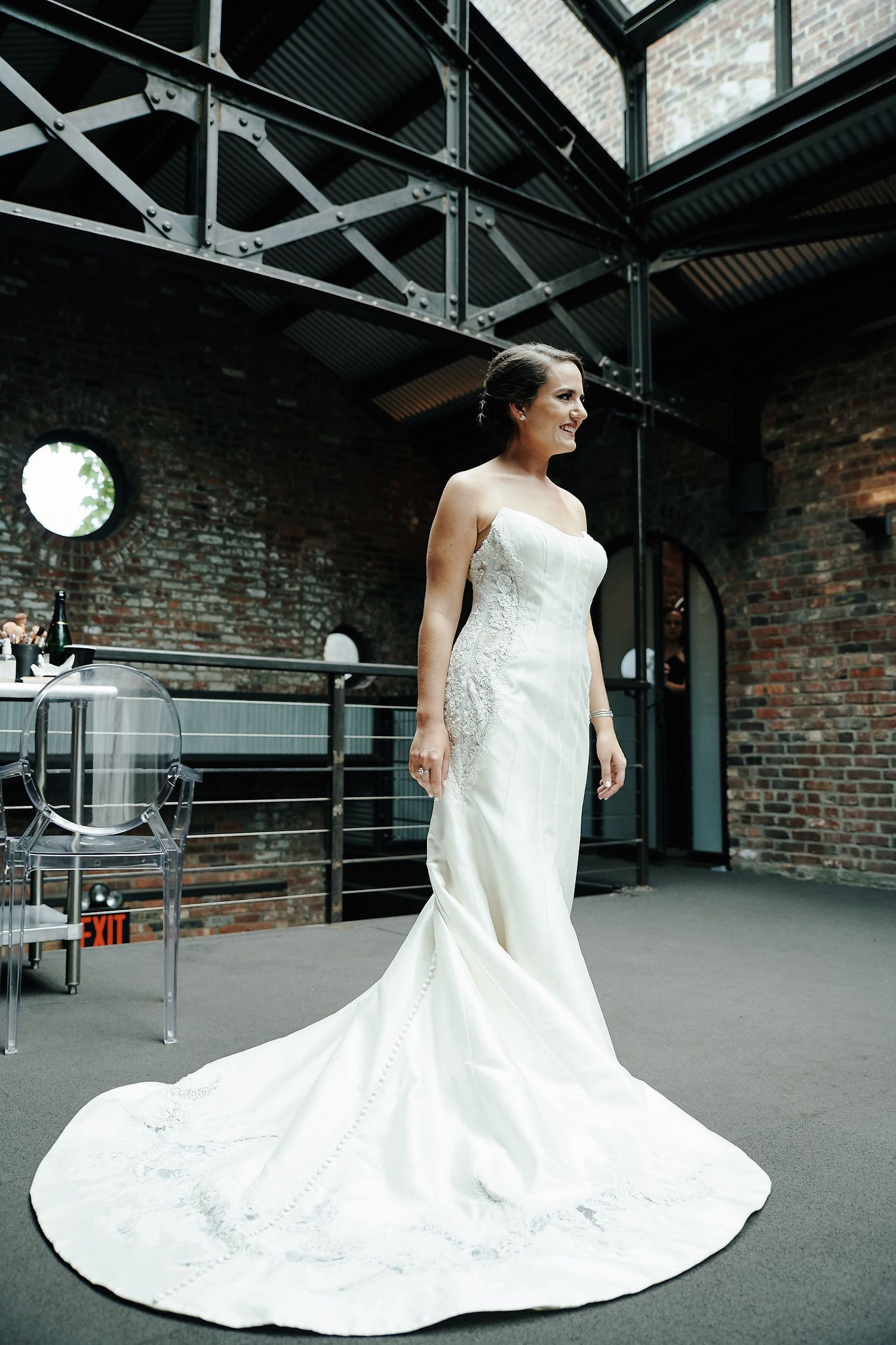 A bride in a fitted white lace gown with a long train poses in an industrial brick venue with skylight.