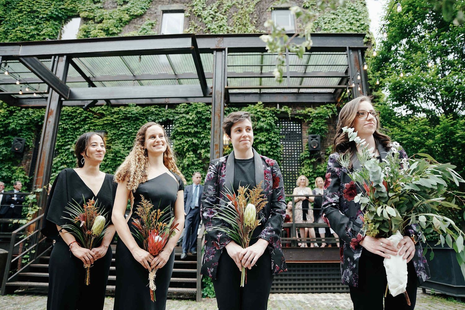 A wedding party stands together holding floral bouquets under an outdoor covered pergola in a garden setting.