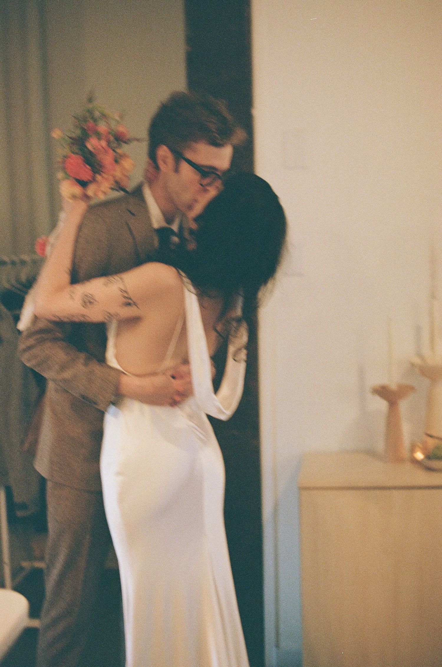 A tender moment between newlyweds dancing in formal attire against a simple indoor backdrop.