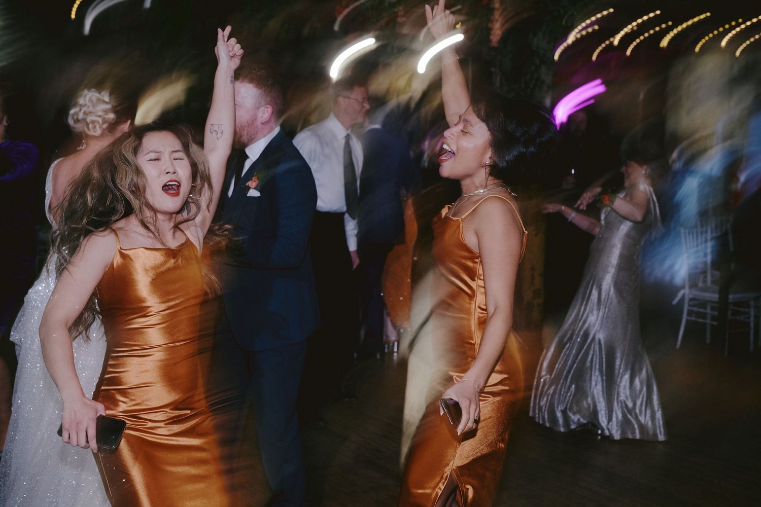 Party guests in shimmering gold dresses dance energetically on a dimly lit dance floor during an evening celebration.