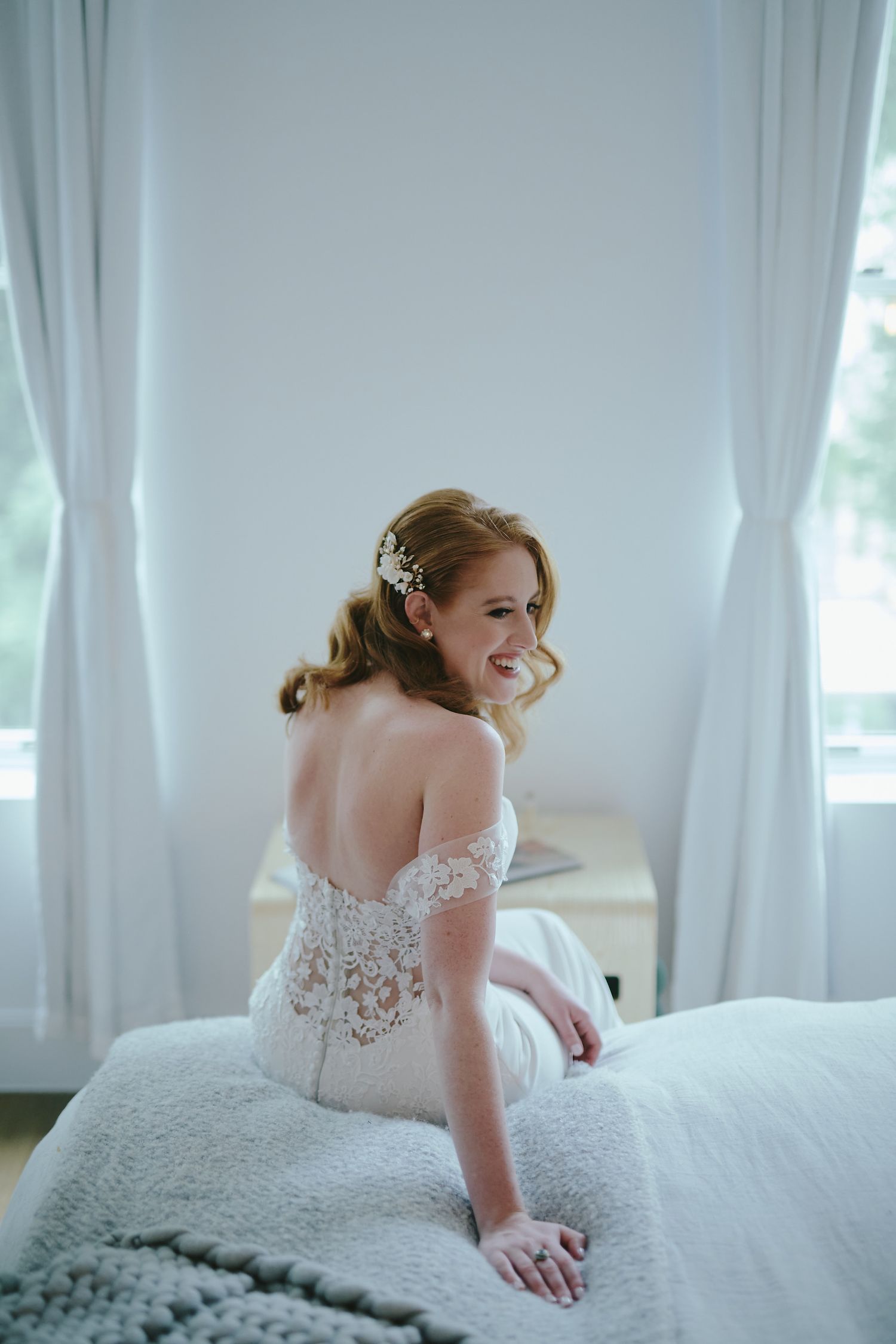 A bride in a delicate off-shoulder lace wedding dress sits on a bed near white curtains in a bright room.