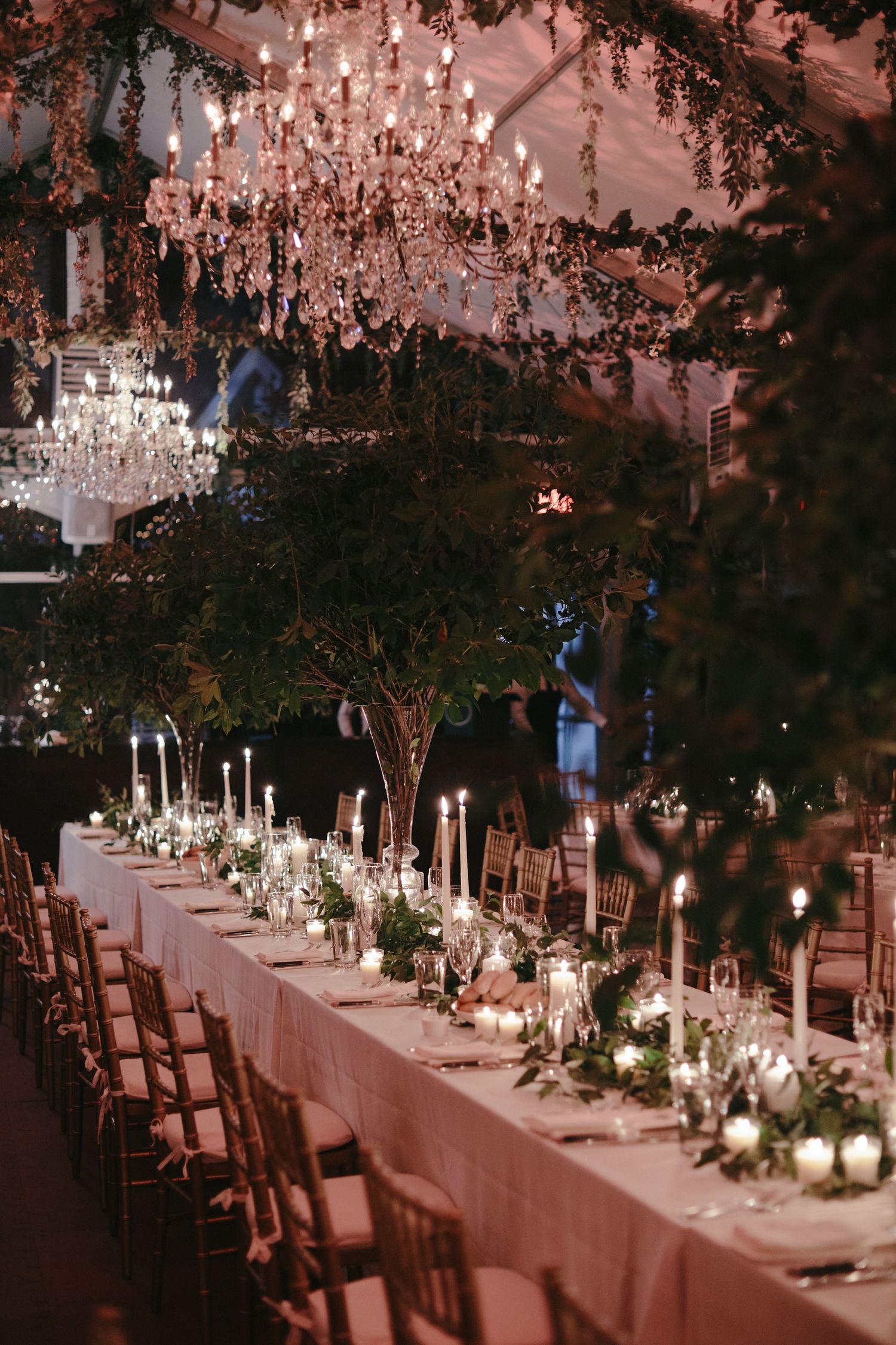 An elegant wedding reception table set with crystal chandeliers overhead, candlelight and floral arrangements on a long table.