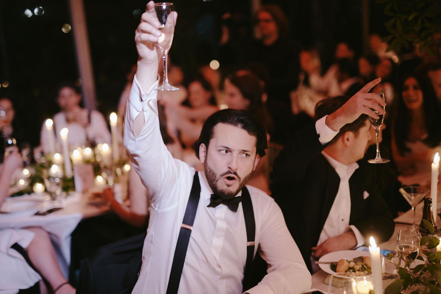 A guest in a white dress shirt and suspenders raises their arms enthusiastically during an elegant candlelit reception.