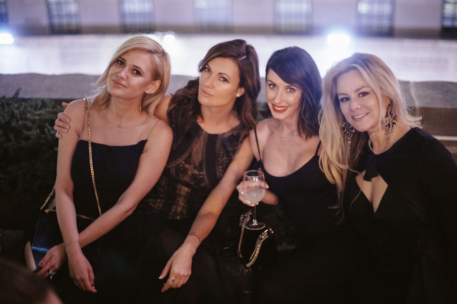 A group of four friends dressed in black cocktail dresses pose together at an evening event.