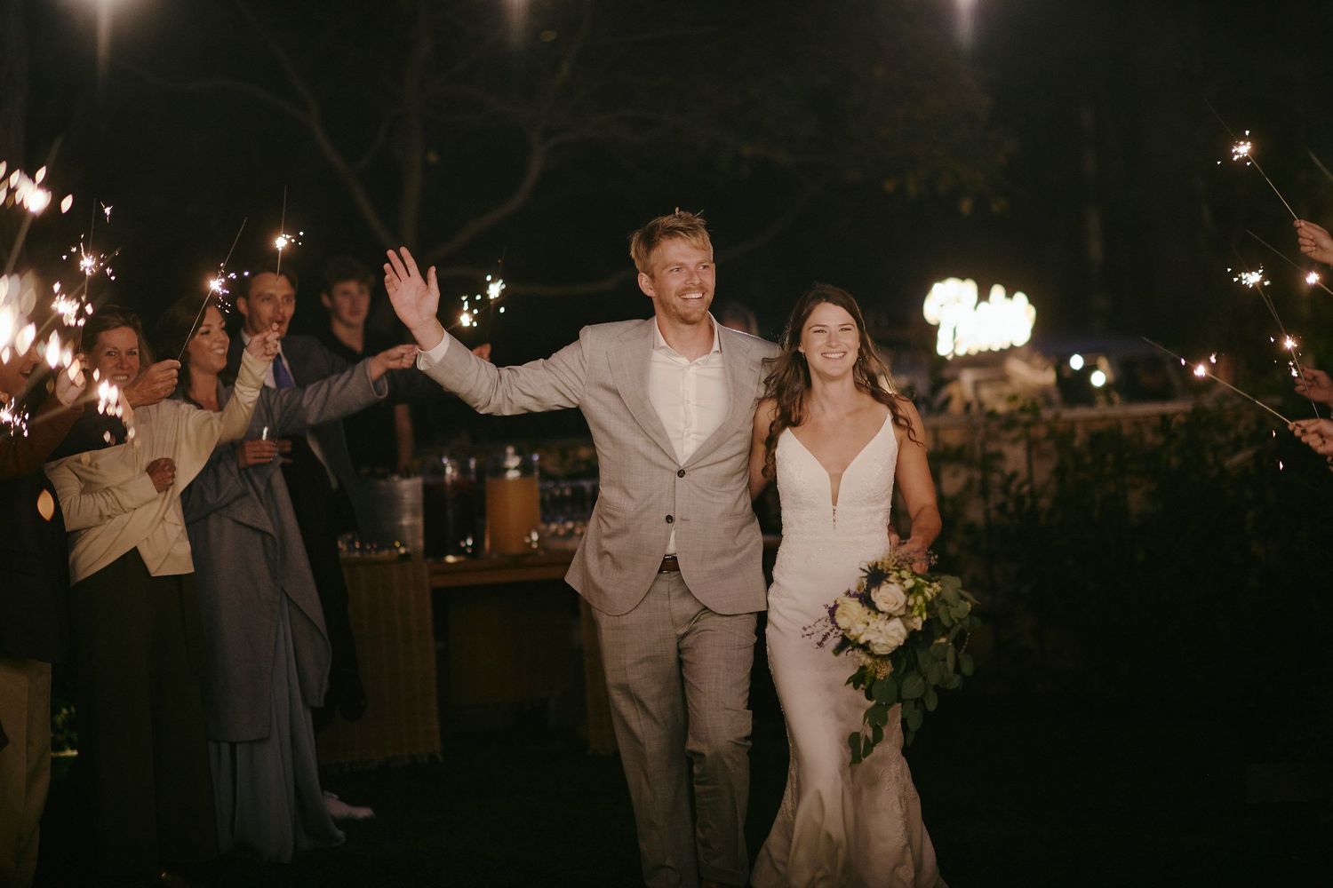 Newlywed couple walks through cheering guests during nighttime wedding celebration with sparklers lighting the path.