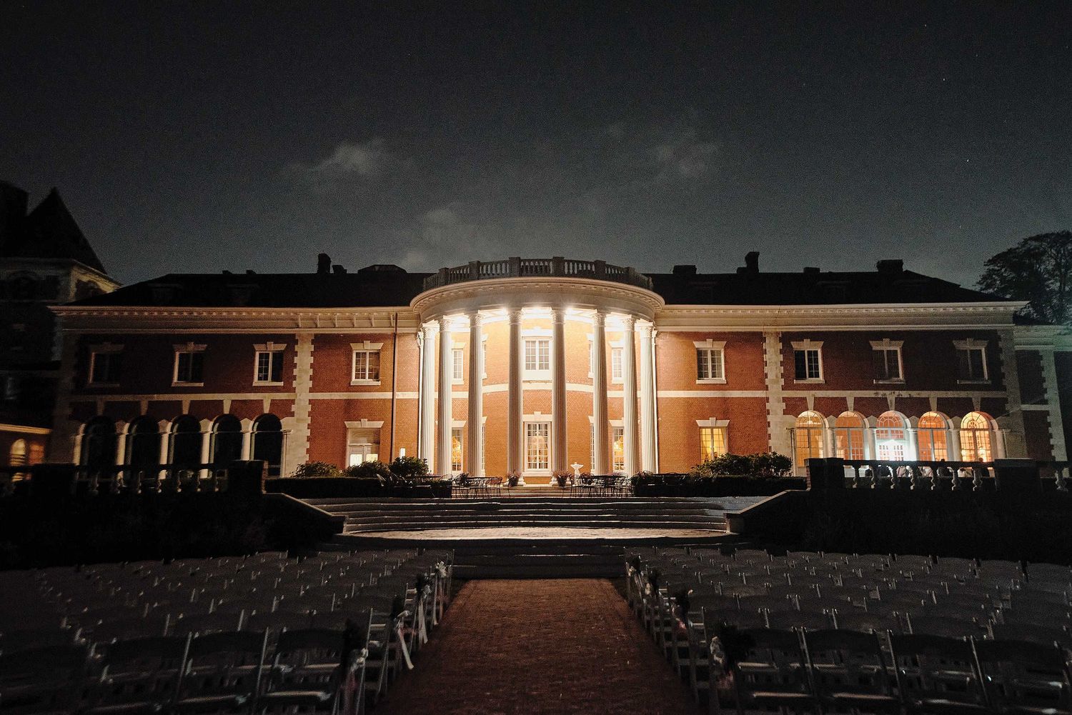 Illuminated neoclassical building with columns at night with rows of empty chairs set up for an outdoor event.
