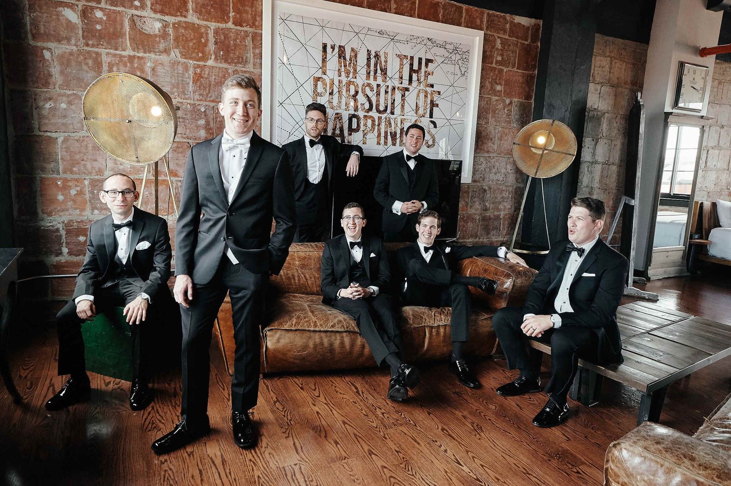 A group of groomsmen in black tuxedos gather around a brown leather couch in an industrial-style loft space.