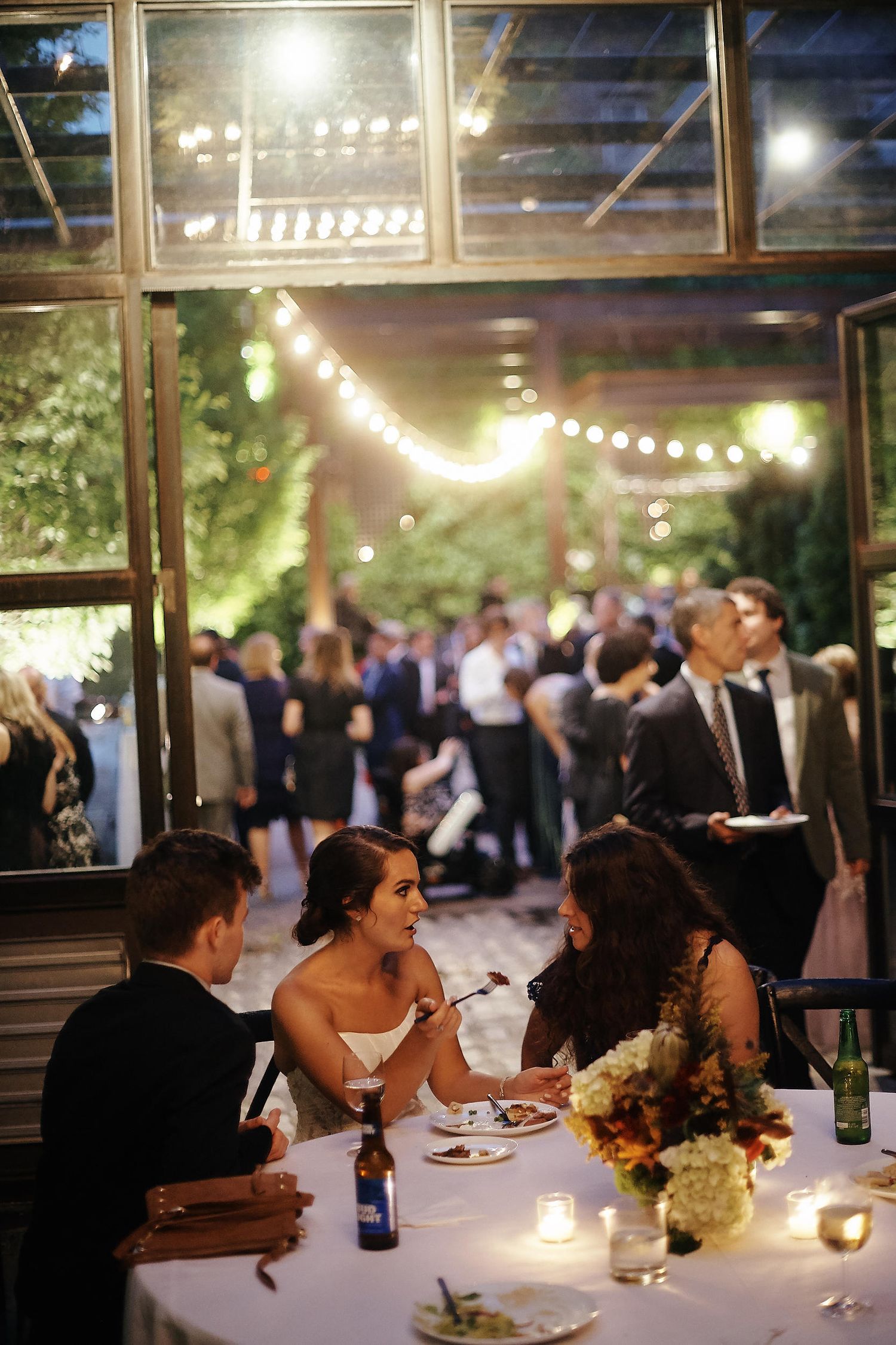 A bride socializes with guests at a candlelit reception table under romantic string lights.