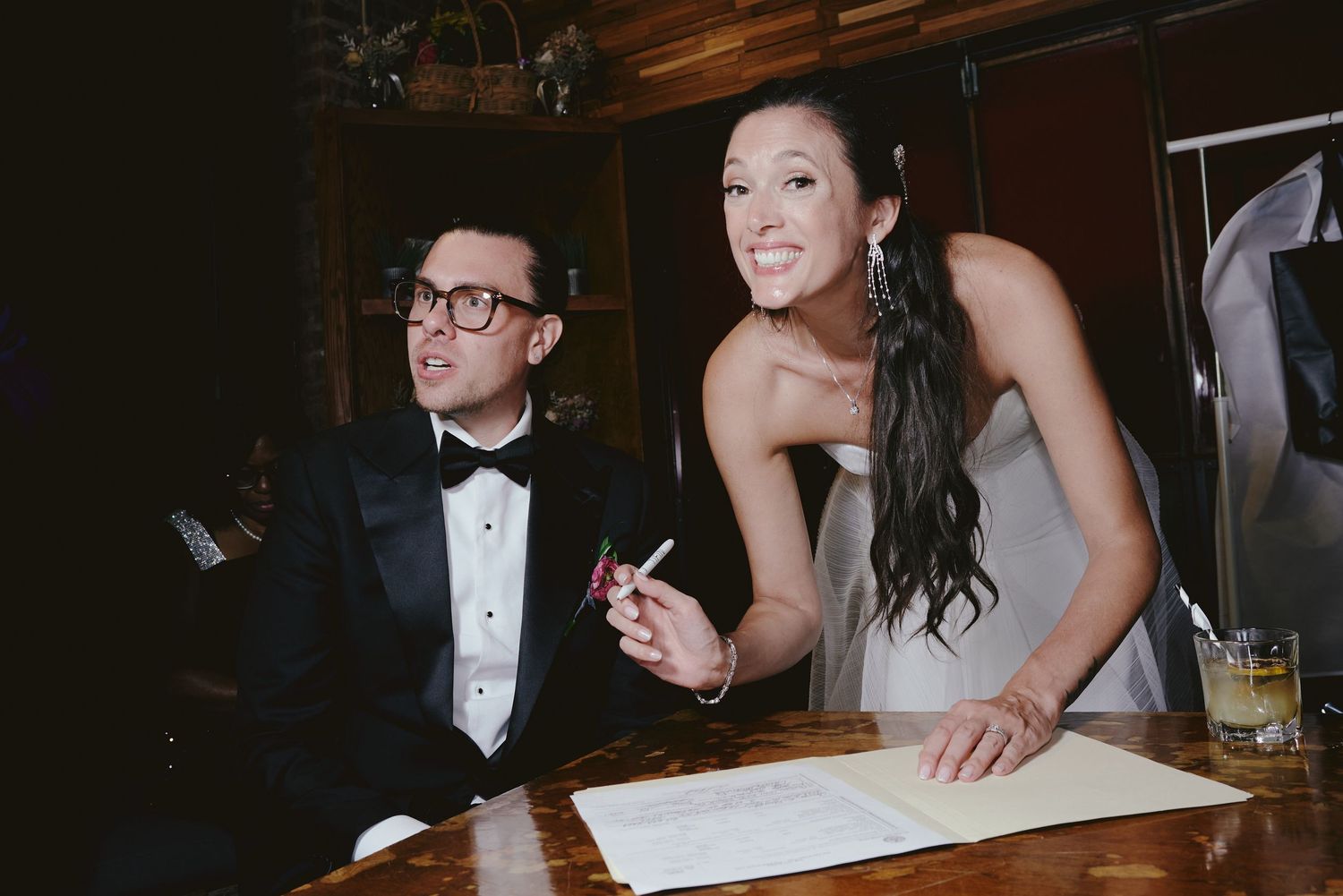Two wedding guests in formalwear lean over a table to sign what appears to be a document at an indoor reception.