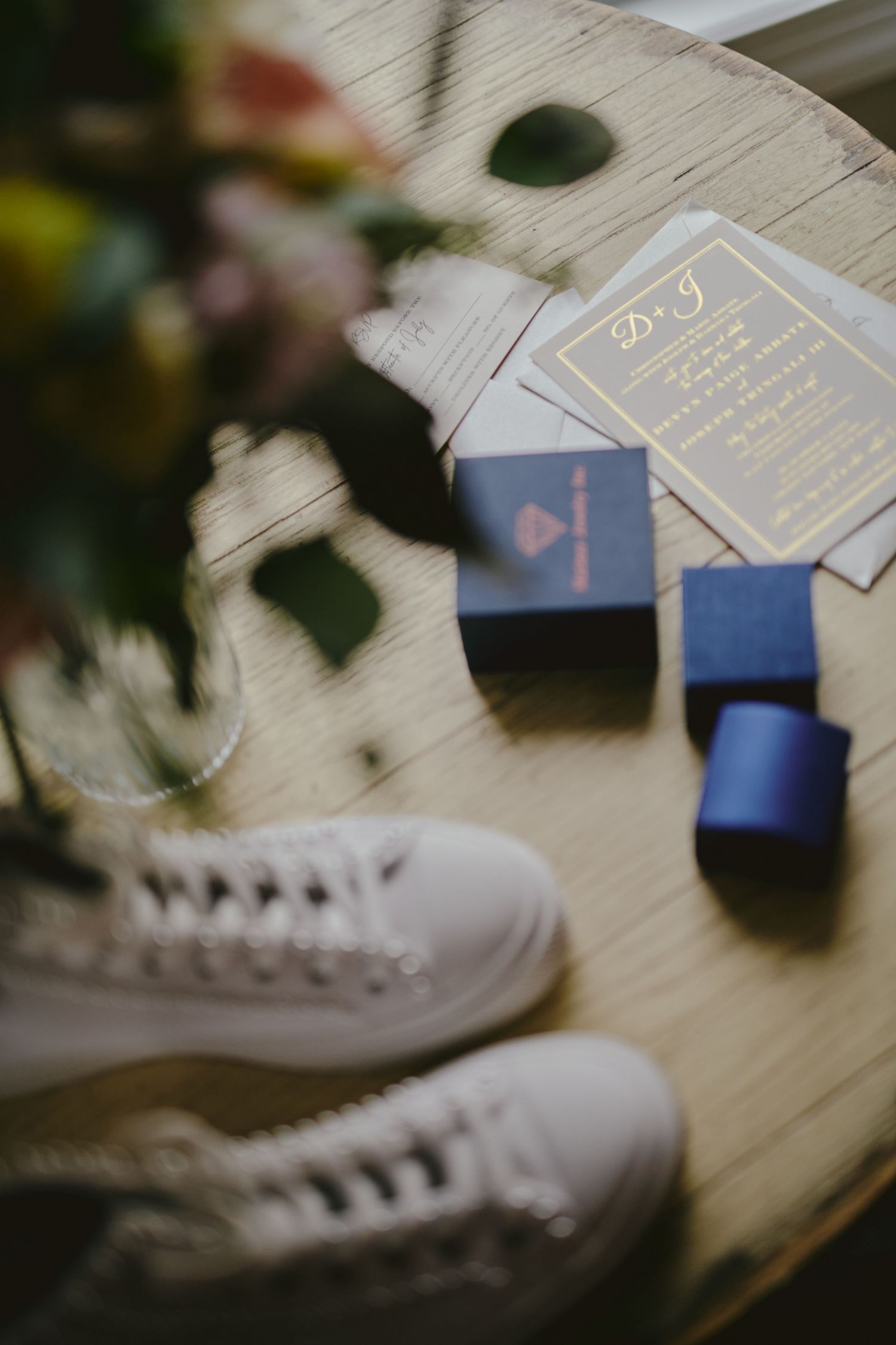 Wedding invitation cards and ring boxes arranged with white sneakers on a light surface next to blurred floral decor.