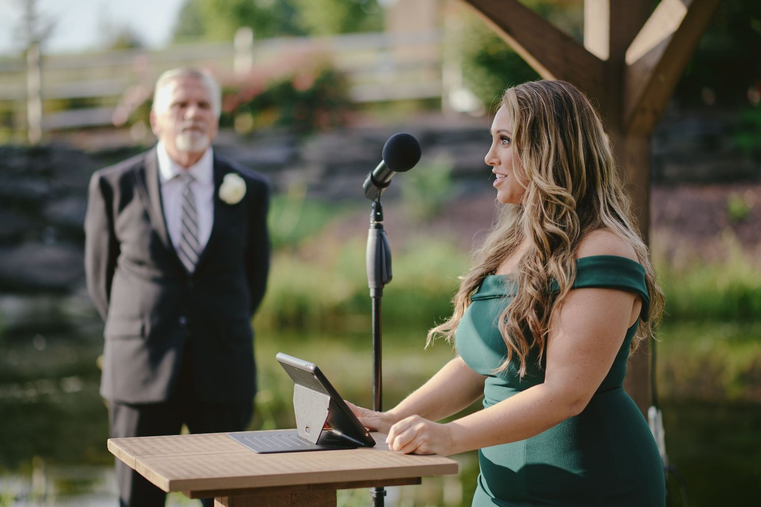 A wedding ceremony speaker stands at a podium outdoors while another individual watches in the background.