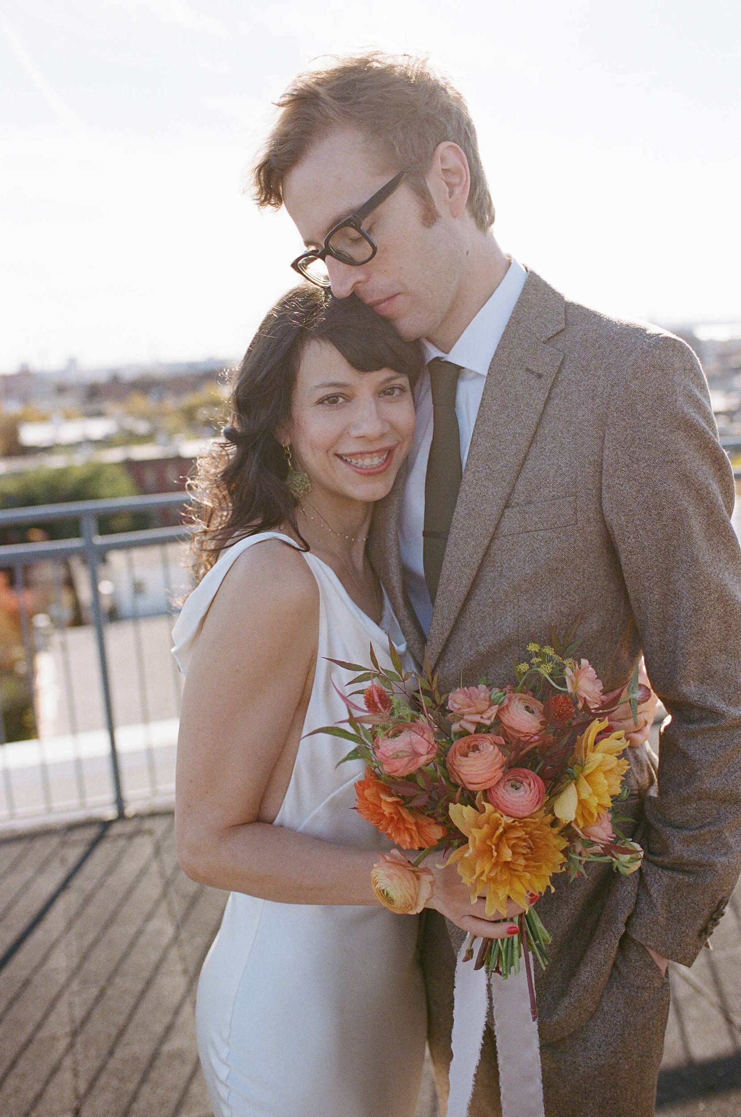 Couple embraces on rooftop terrace at sunset as bride holds vibrant orange and pink floral bouquet.