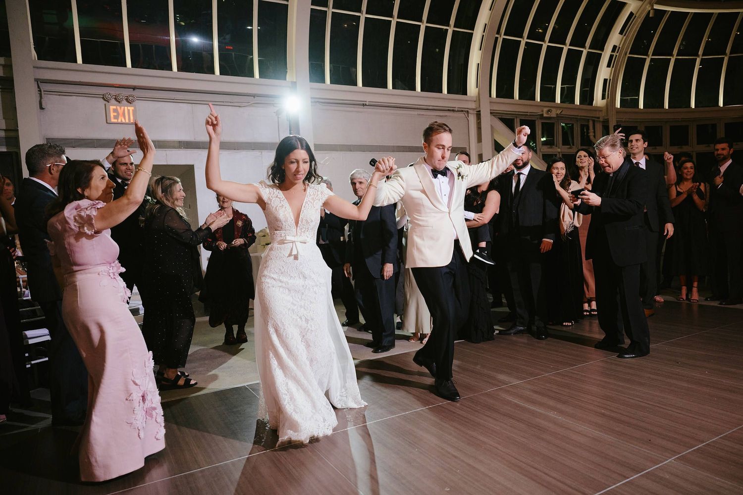 Wedding party dances joyfully during reception celebration in an elegant glass-ceiling ballroom.