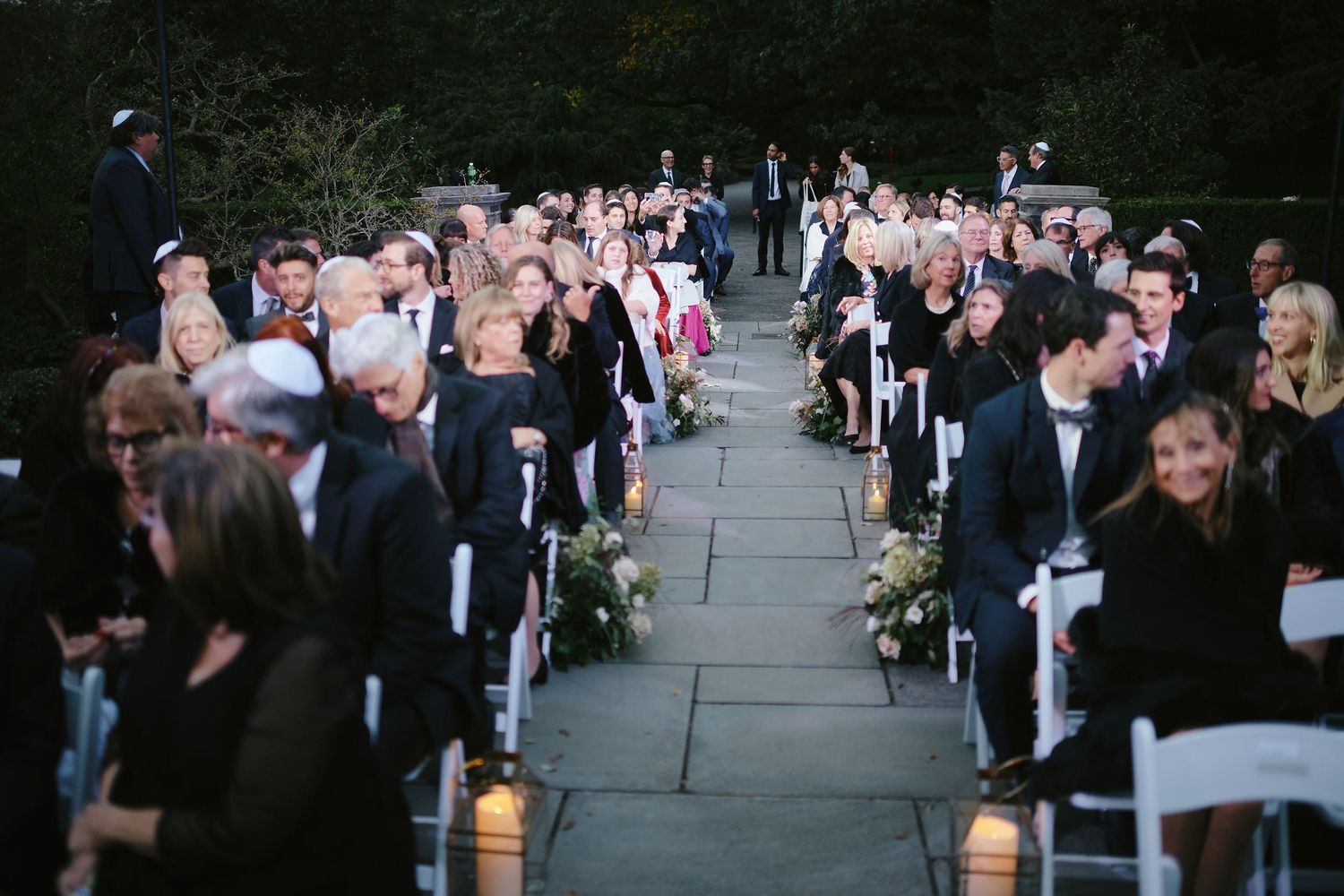Wedding ceremony guests sit in white chairs along a stone pathway lined with lanterns and floral decorations at dusk.