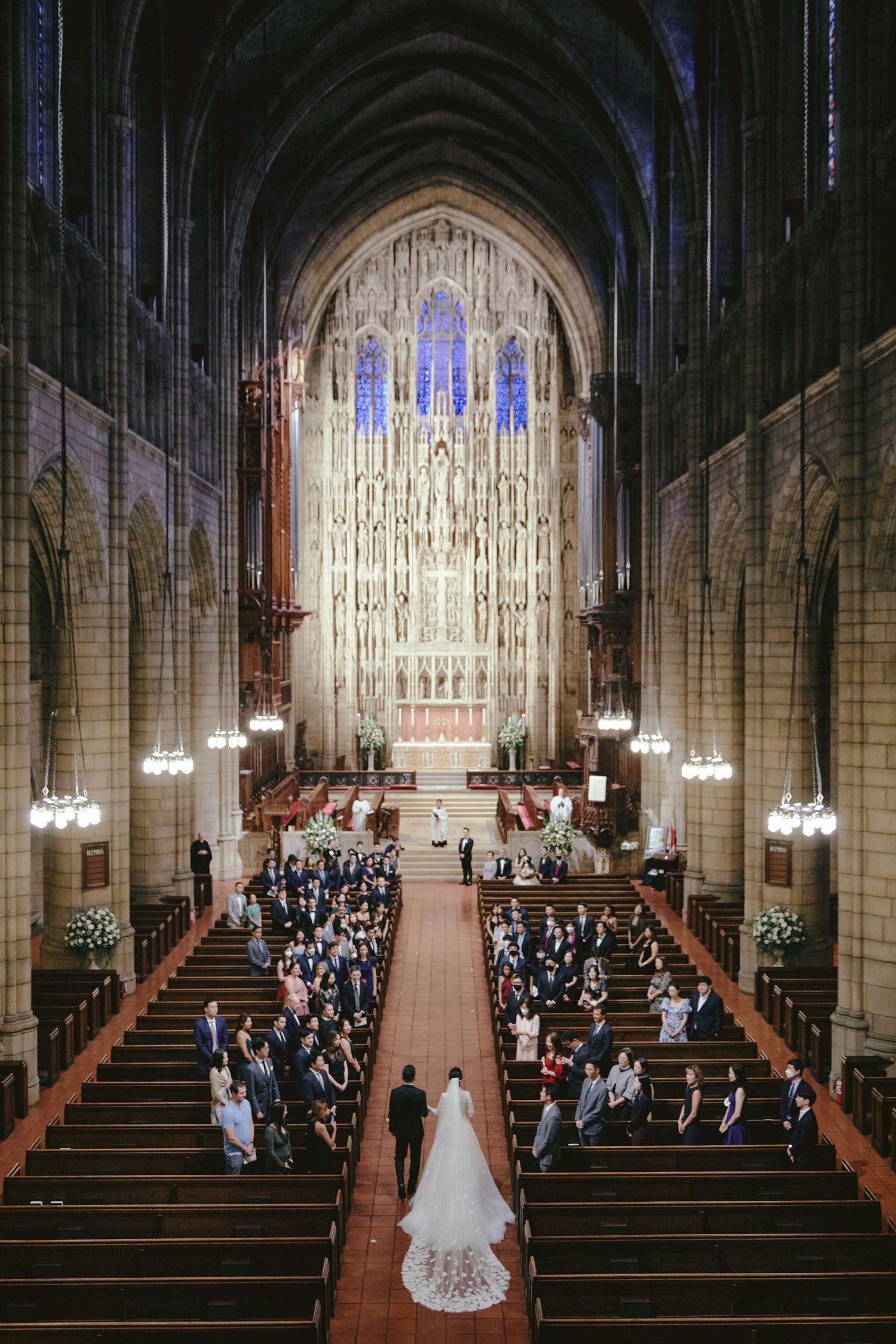 Bride walking down the aisle of a grand Gothic cathedral church during a wedding ceremony with stained glass windows.